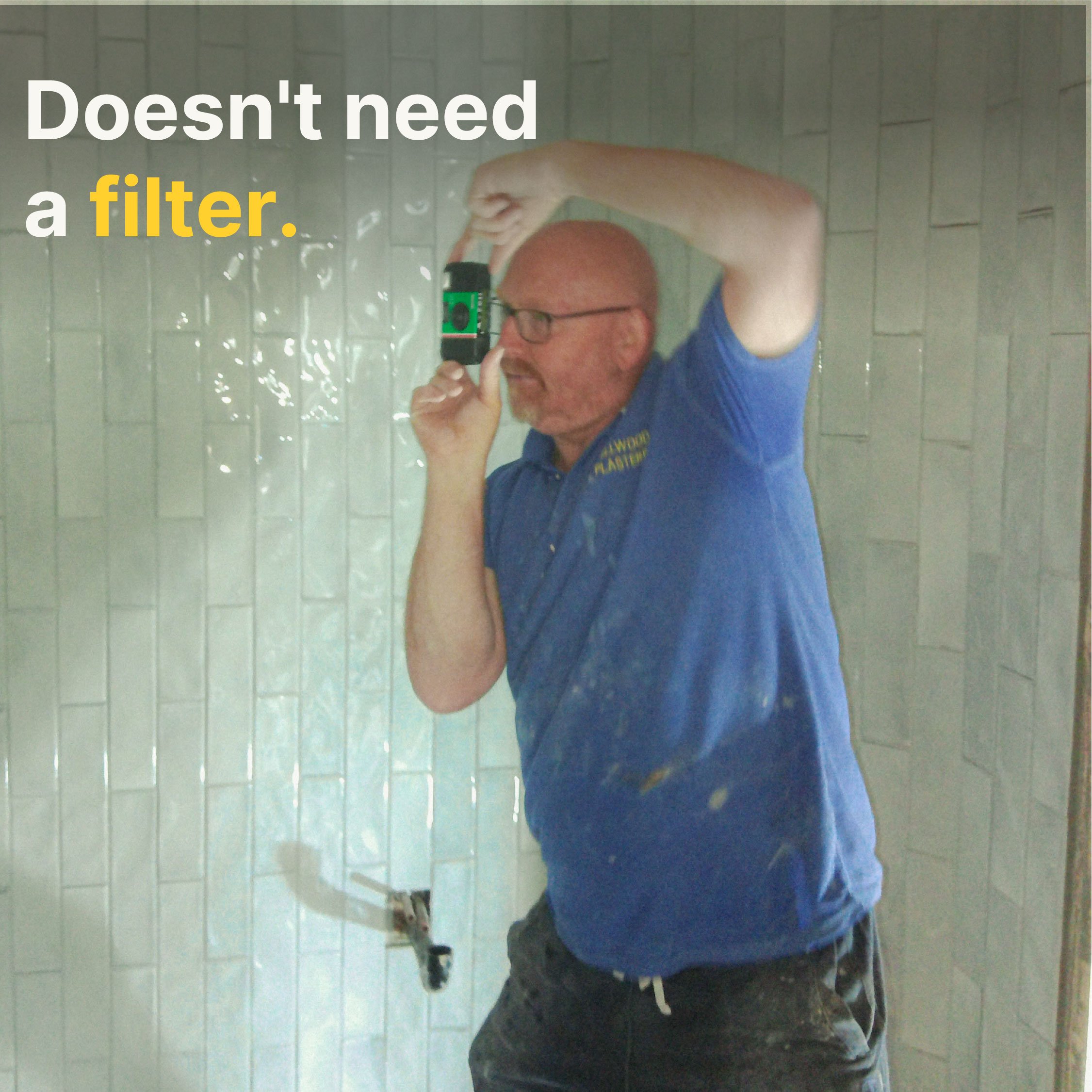 A man with glasses taking a selfie in a mirror in a tiled room, wearing a blue shirt, holding a phone, with a wheelchair in the background.