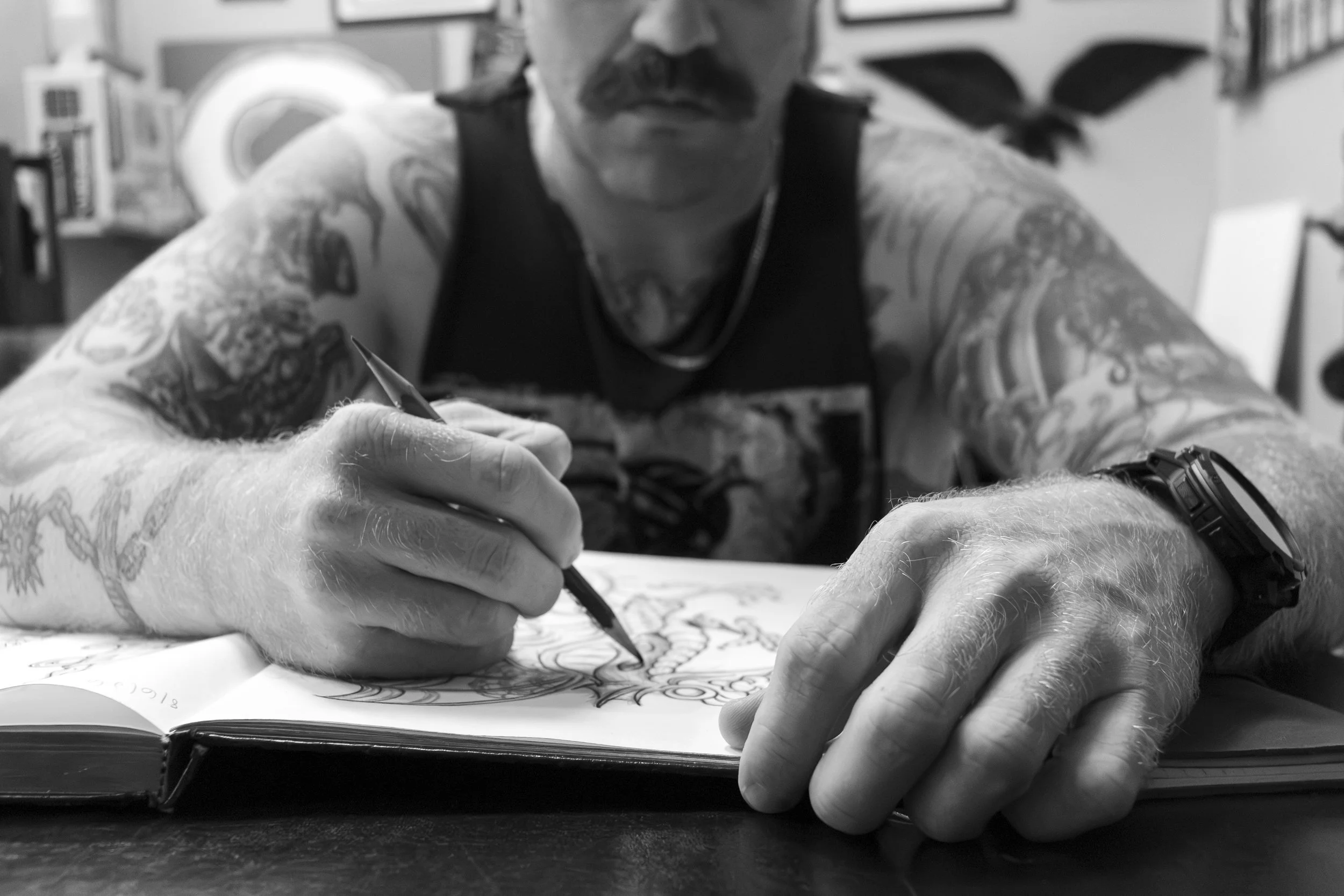 A person with tattoos on their arms is drawing in a sketchbook with a pen. The individual has a mustache, is wearing a tank top, and is sitting at a table.