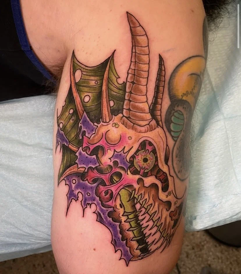 Colorful tattoo of a skull with dragon-like features, including horns and spiky teeth, on a person's upper arm.