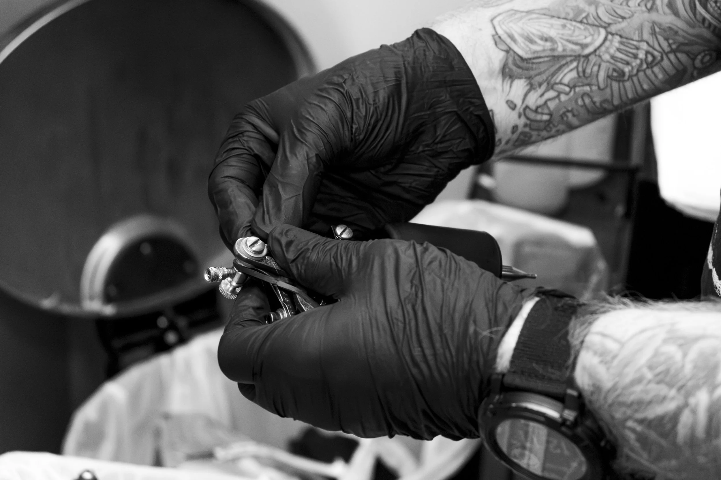 A tattooed person wearing black gloves is tattooing someone with a tattoo machine. The image is in black and white.