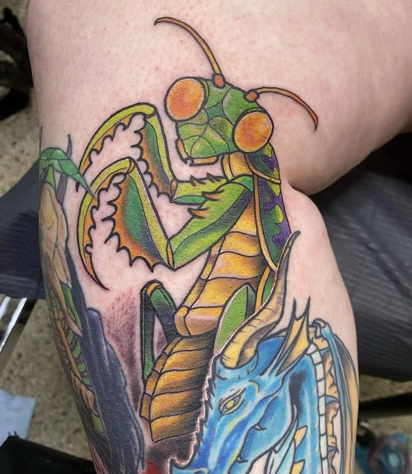 Colorful tattoo of a praying mantis, a dragon, and a blue salamander on a person's thigh.