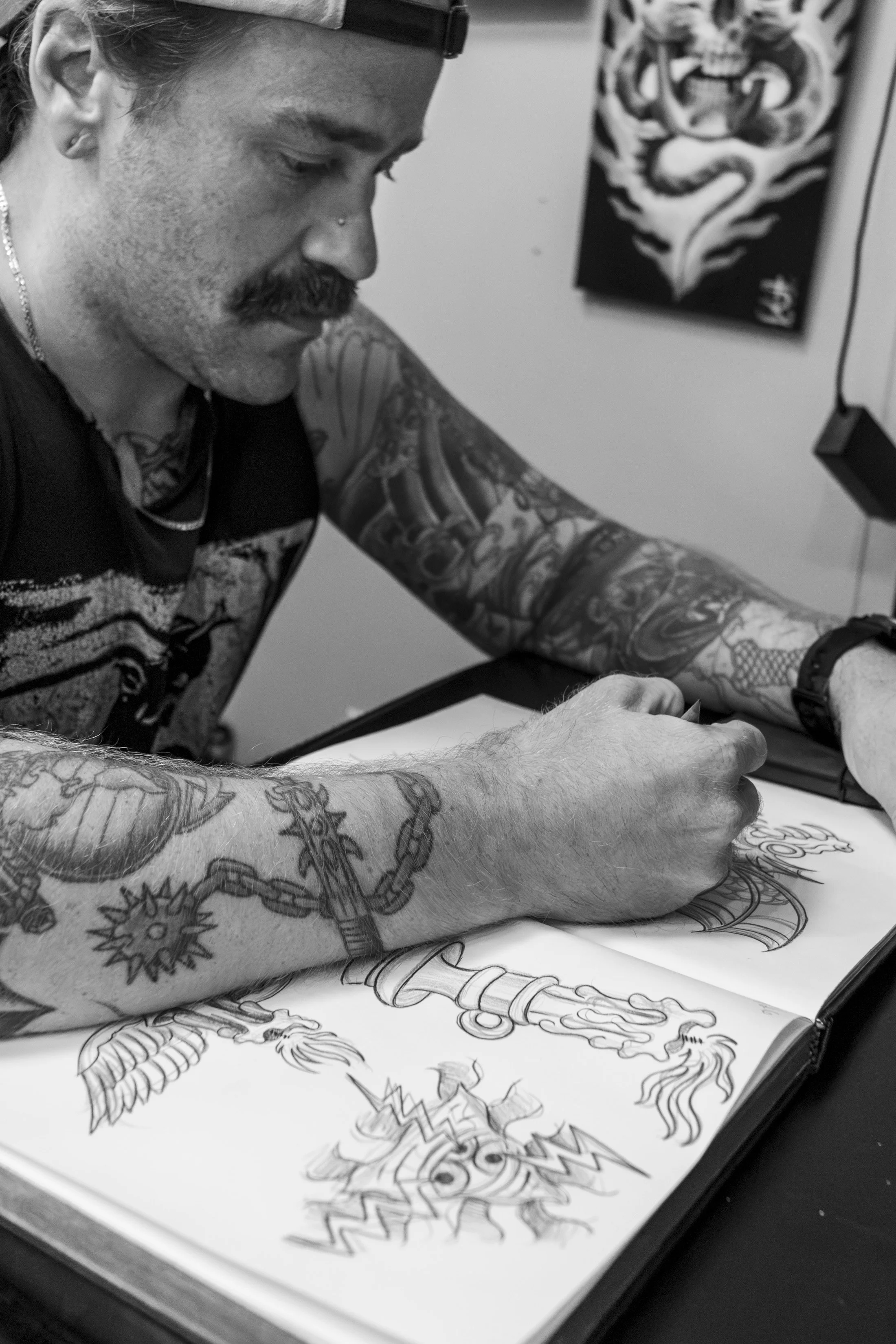 A person with tattoos on their arms and a mustache, wearing a cap, is drawing or inking an intricate dragon design on a large sheet of paper.