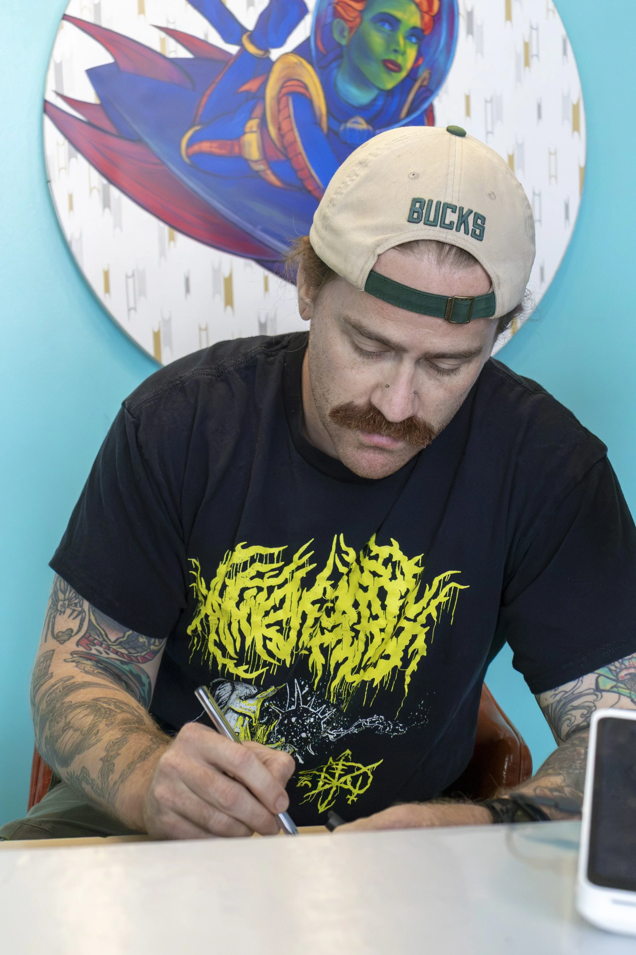 A man with a beard, tattoos on his arms, and wearing a beige cap with the word 'BUCKS' on it is seated at a table, writing on paper with a pen. Behind him, there is colorful wall art depicting a woman with vibrant makeup, green skin, and a superhero costume, set against a teal wall.