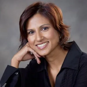 A woman with short, reddish hair smiling and resting her chin on her hand, wearing a black blazer against a gray background.