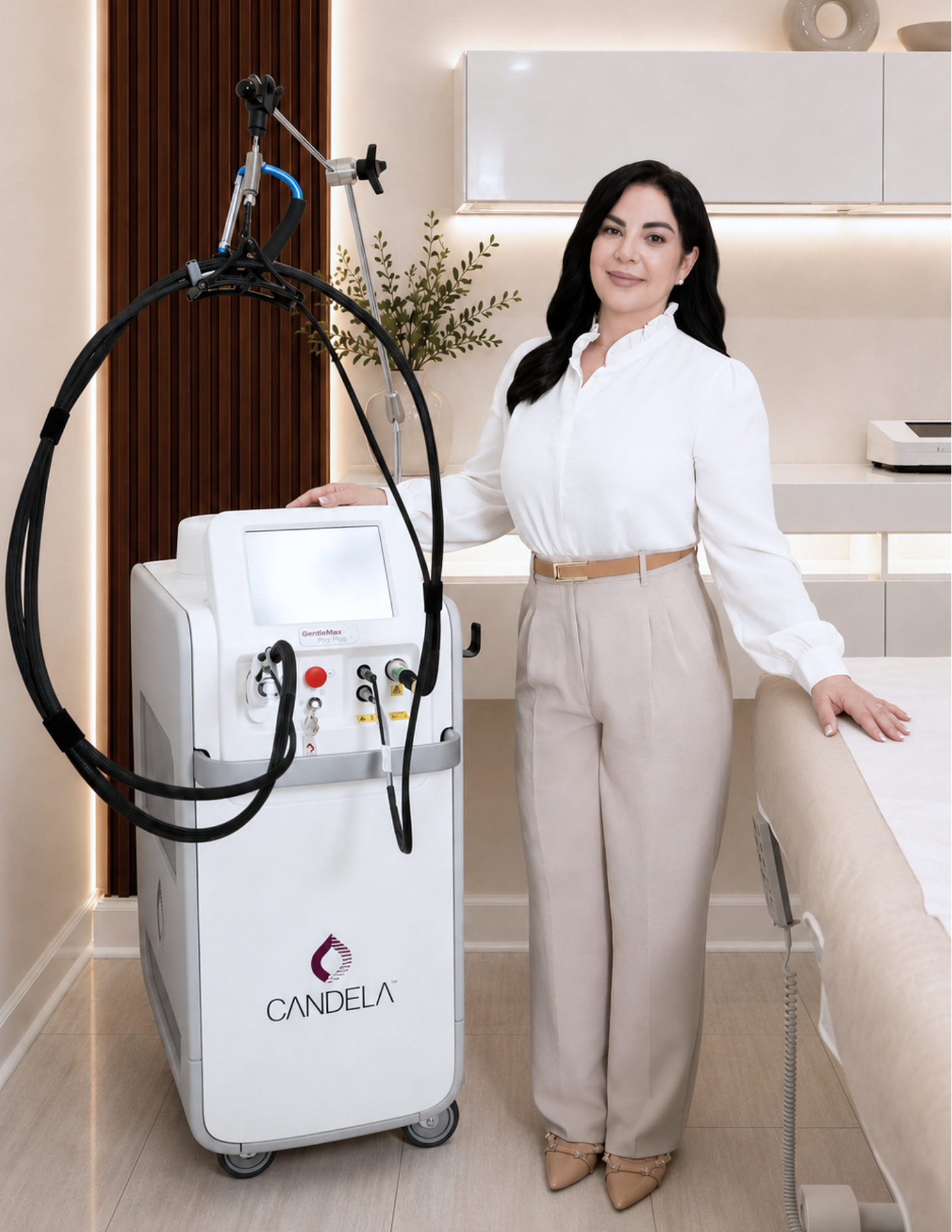 A woman with black hair and a white blouse standing in a medical or spa setting, next to a white machine with a screen and hoses, with the logo 'CANDELA' on it. There is a bed or examination table to her right and decorative items in the background.