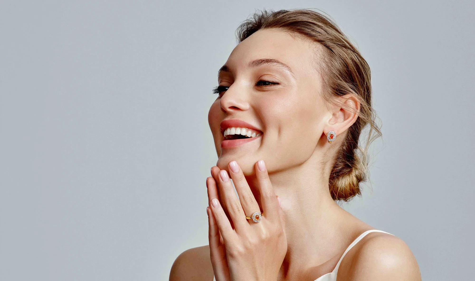 A smiling woman with light skin, wearing earrings, ring, and a necklace, touching her chin with her fingers, against a plain gray background.
