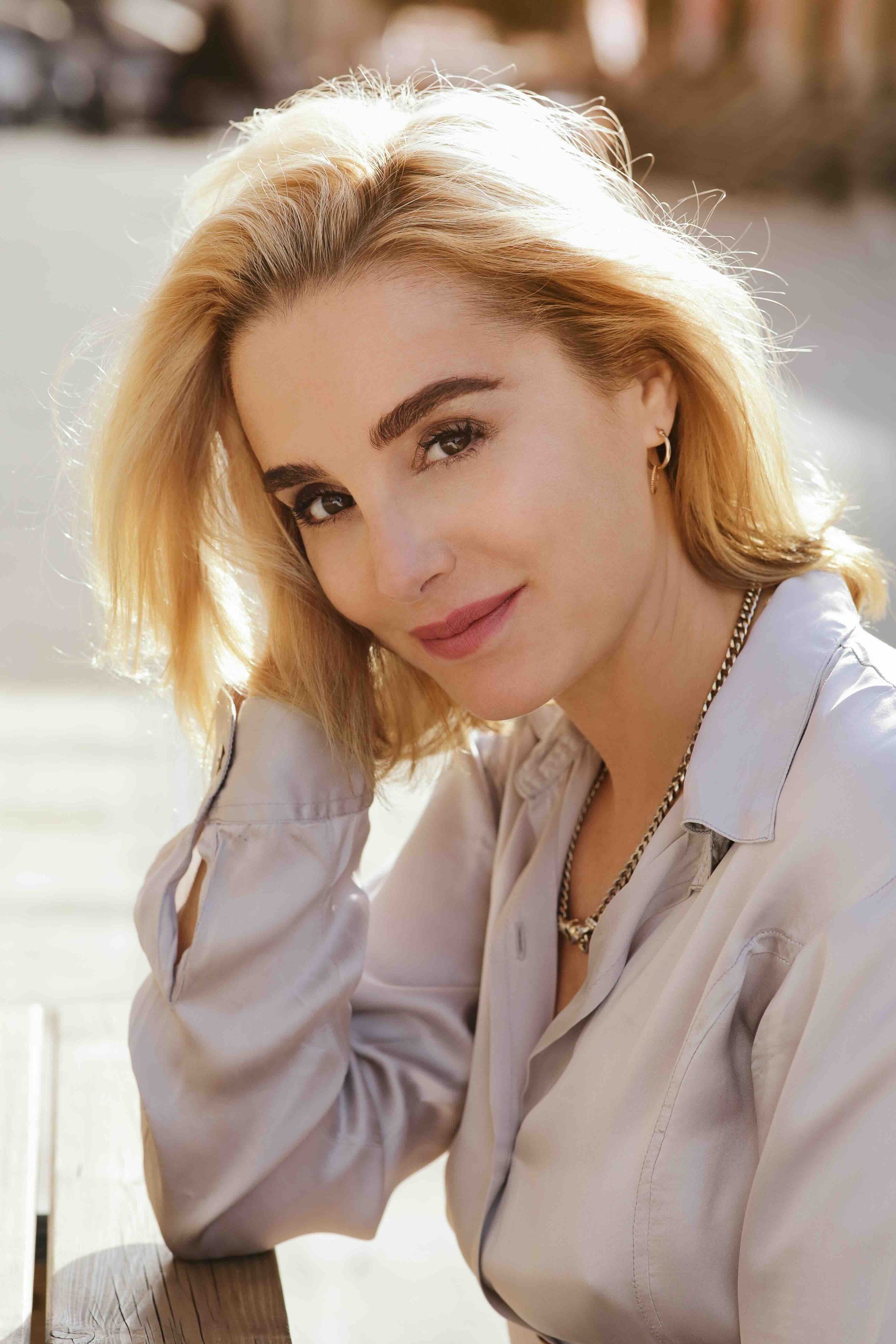 Close-up of a woman with blonde hair, wearing a beige shirt and jewelry, smiling softly at the camera outdoors with sunlight on her face.