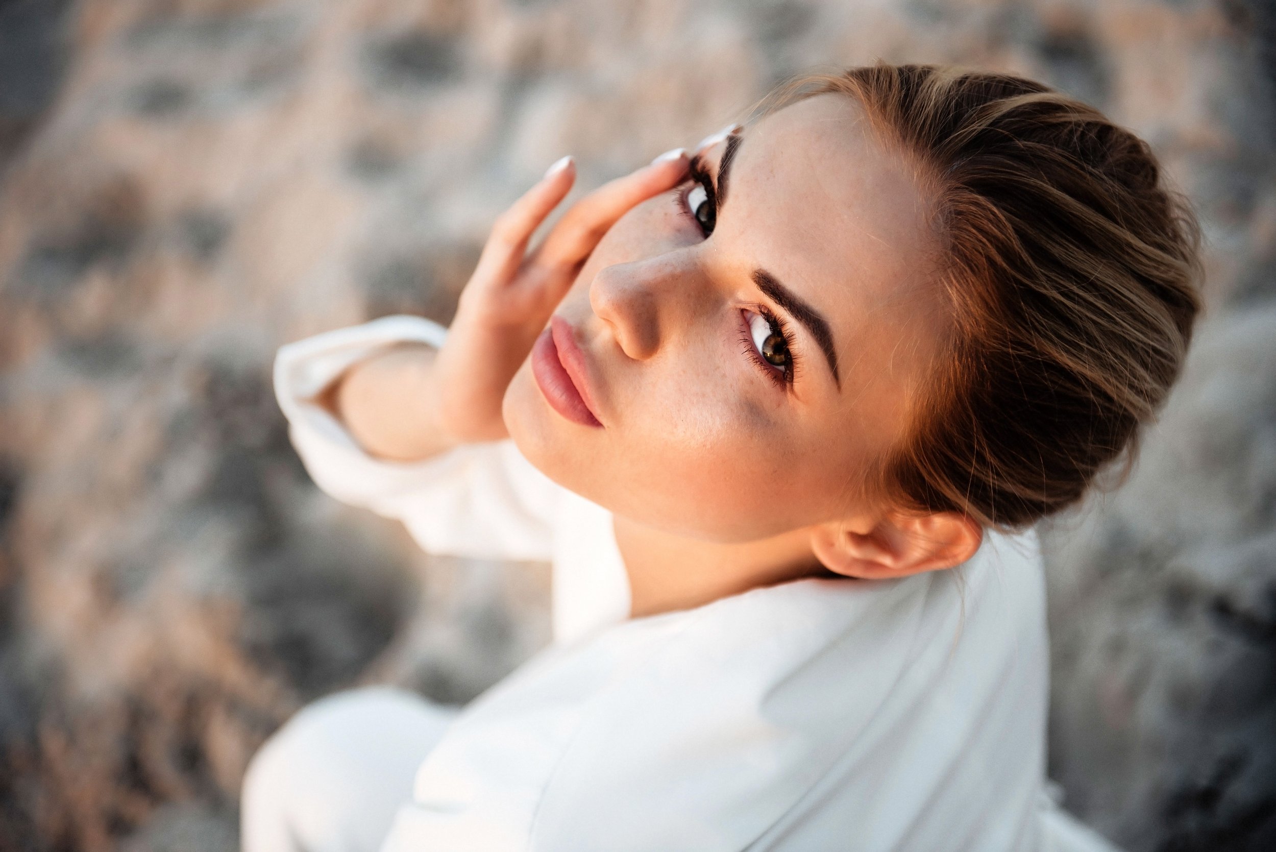 Close-up of a woman with brown hair and brown eyes, looking up at the camera with her hand near her forehead, outdoors with blurred earth tones background.