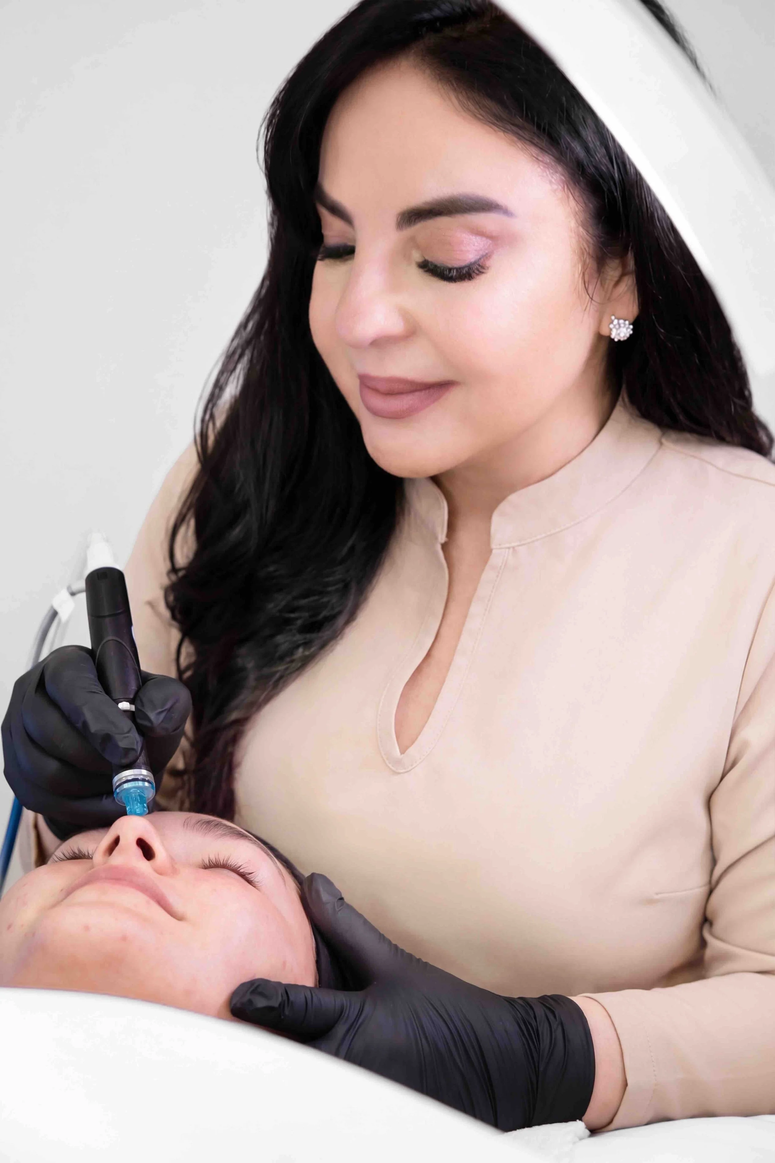 A woman with dark hair and earrings is holding a syringe close to a patient’s face during a cosmetic procedure, with the patient lying down with closed eyes.