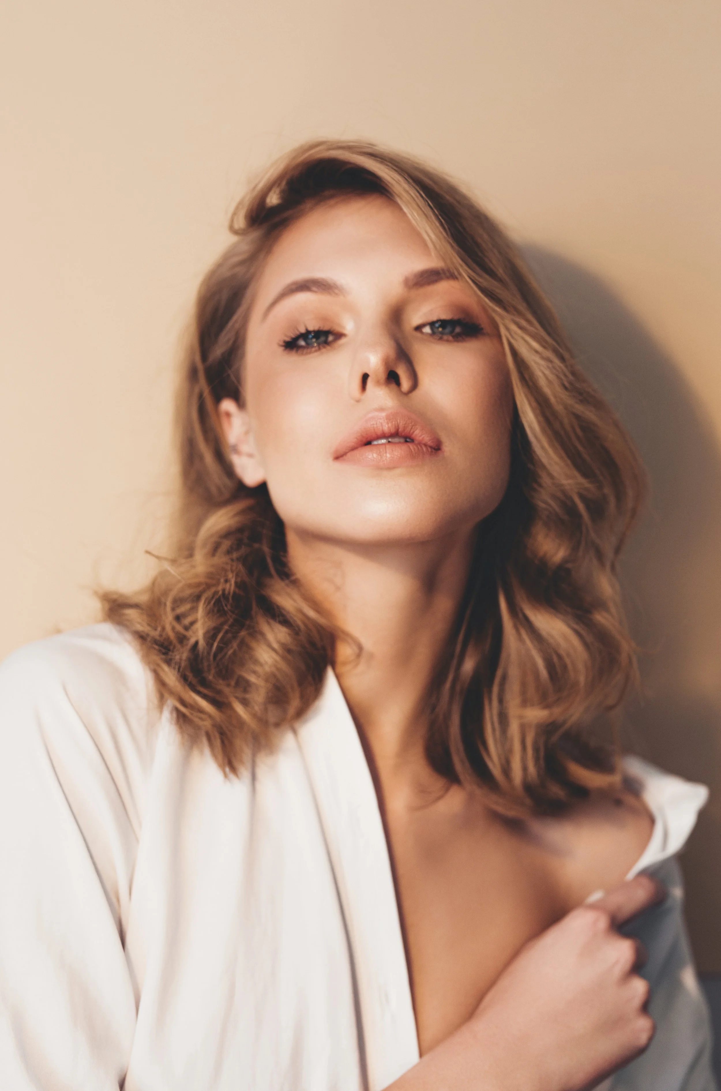 A woman with wavy, shoulder-length brown hair and light makeup, wearing a white satin robe, looking confidently at the camera against a plain beige background.