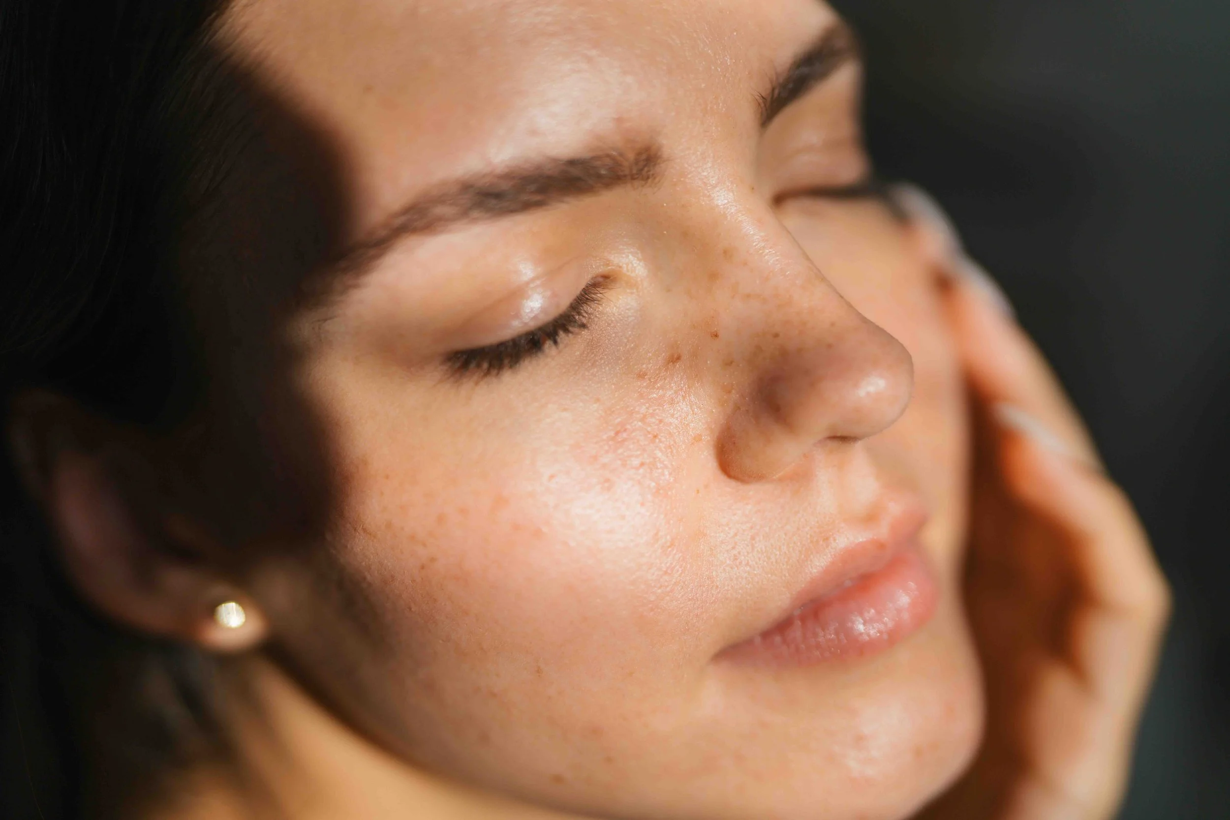Close-up of a woman's face with closed eyes, showcasing clear skin, freckles, and dark hair.