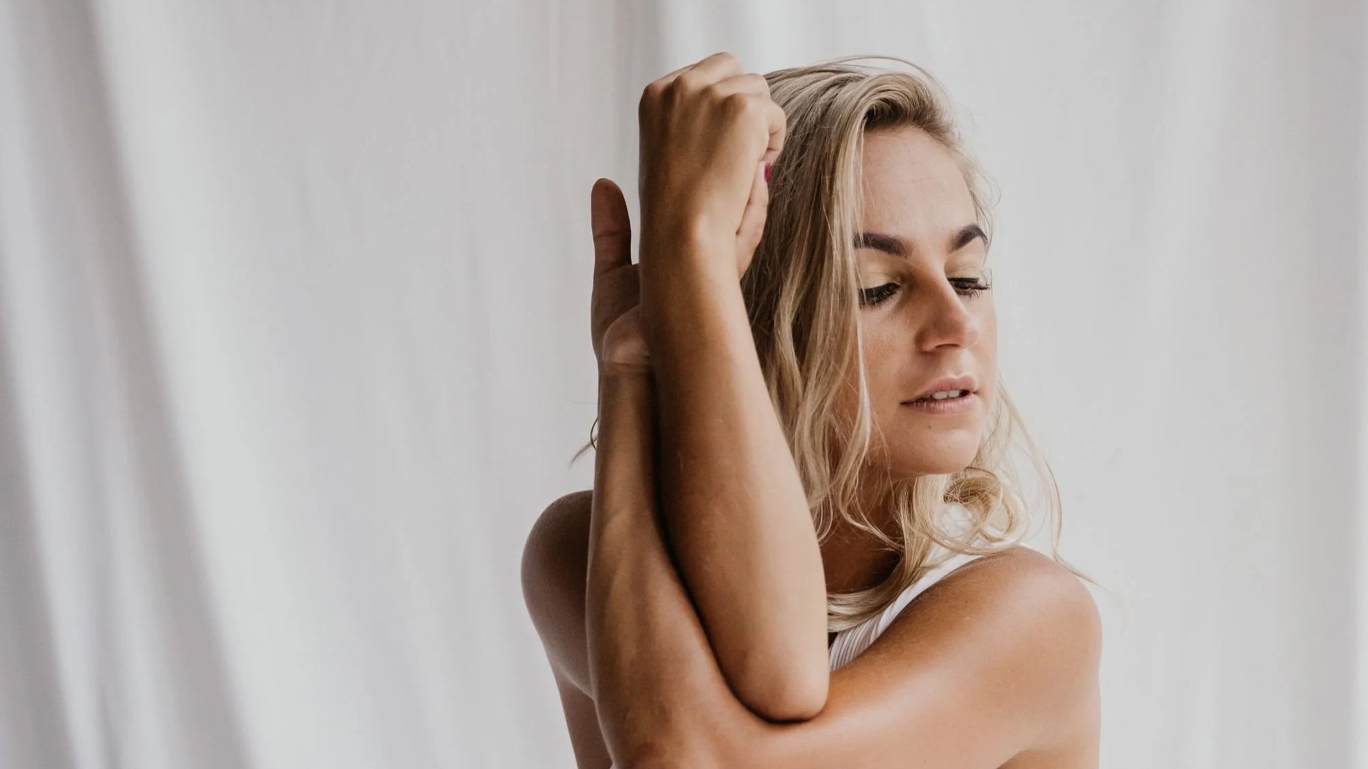 A woman with blonde hair and light skin is relaxing with her eyes closed, while another arm wraps around her shoulder and gently touches her forehead.