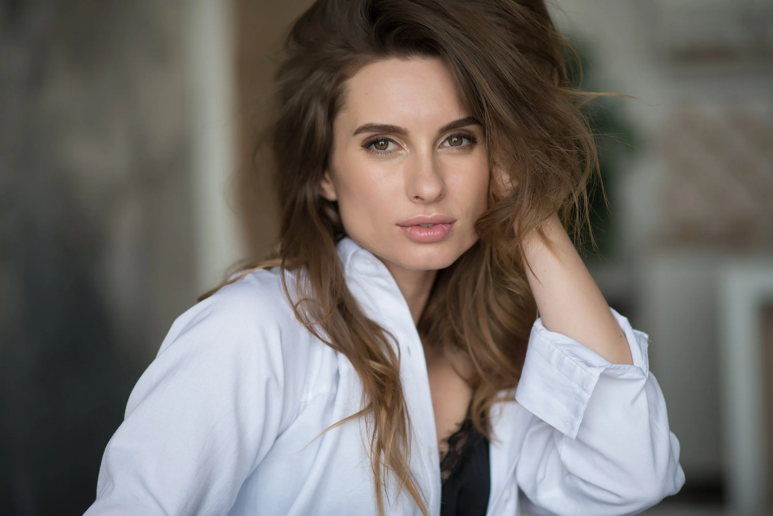 Close-up of a woman with wavy brown hair, olive skin, and green eyes, wearing a white shirt, resting her head on her hand, with a blurred background.