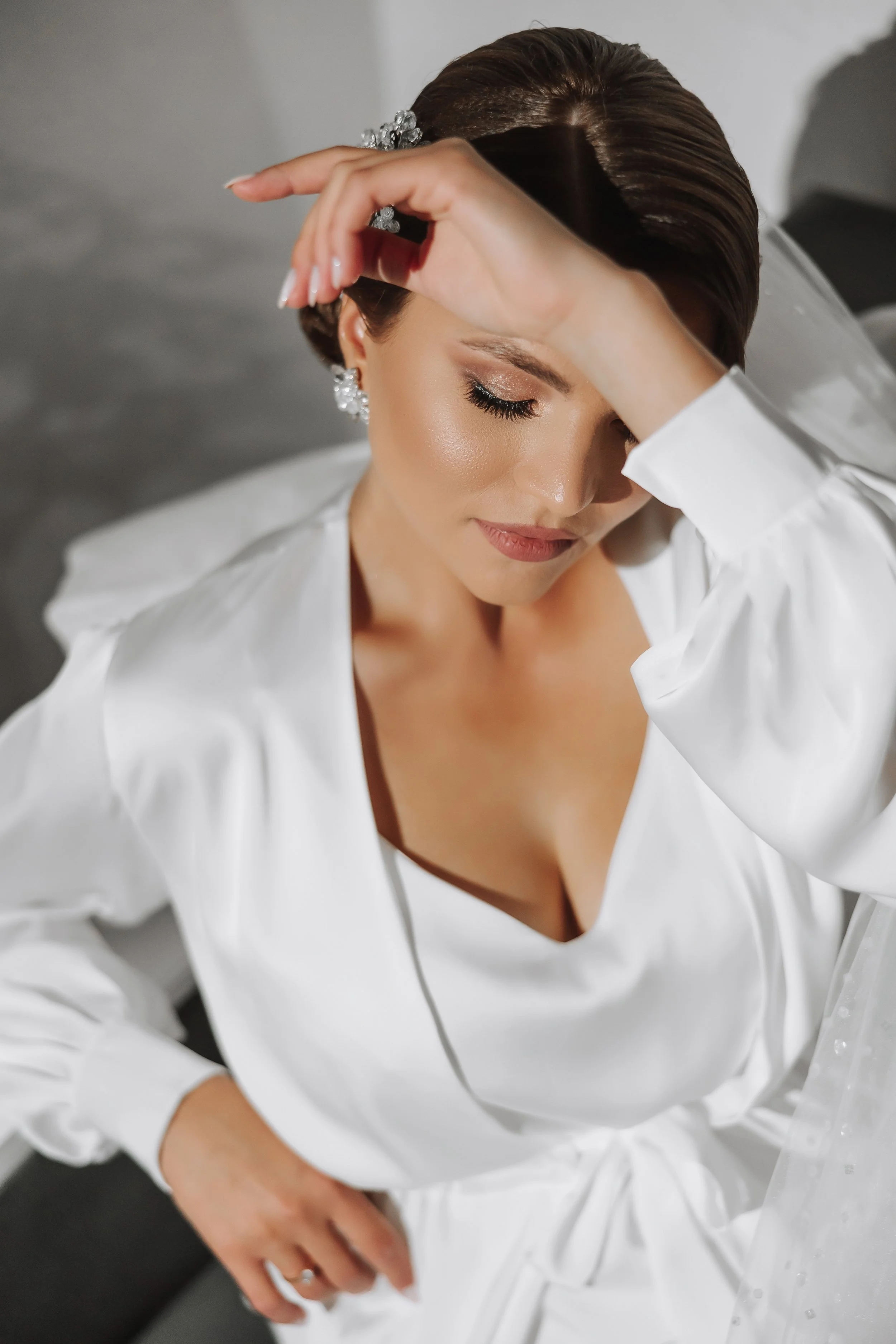 Close-up of a woman with styled brown hair, closed eyes, and makeup, wearing diamond earrings and a white silky shirt, with her hand resting on her forehead.