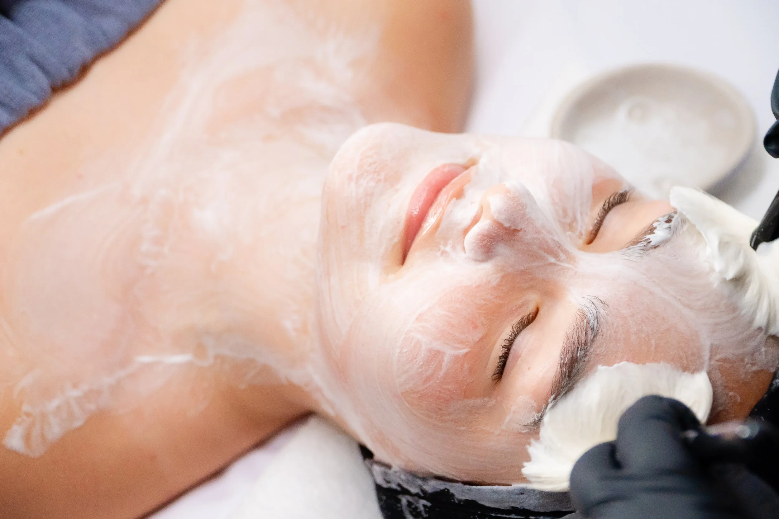 Person lying down while receiving a facial treatment, with cream applied to their face, and a therapist or esthetician working on their skin.