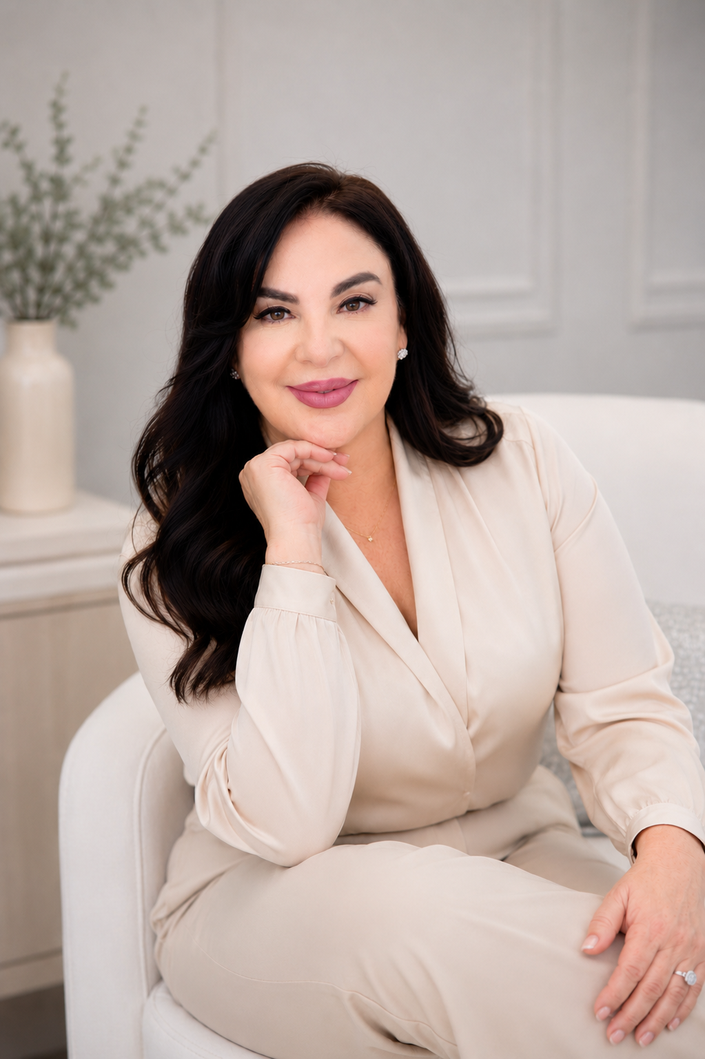 A woman with long dark hair and a warm smile, wearing a cream-colored blouse, sitting on a white chair in a light, elegant room with a vase of greenery in the background.