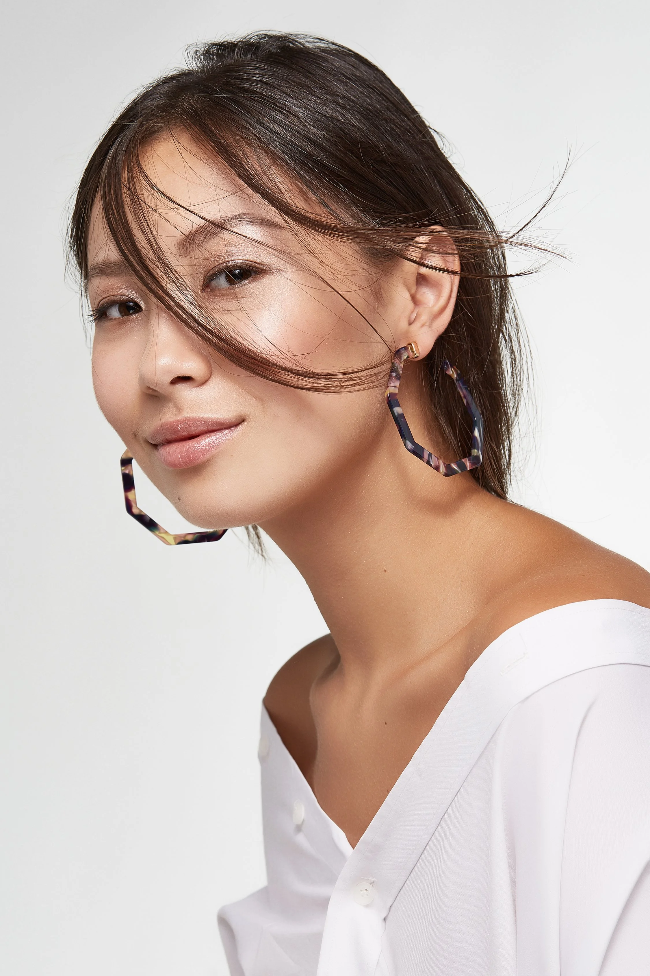 A woman with short brown hair wearing large, dark, marbled hoop earrings and a white button-up shirt, posing against a plain light background.