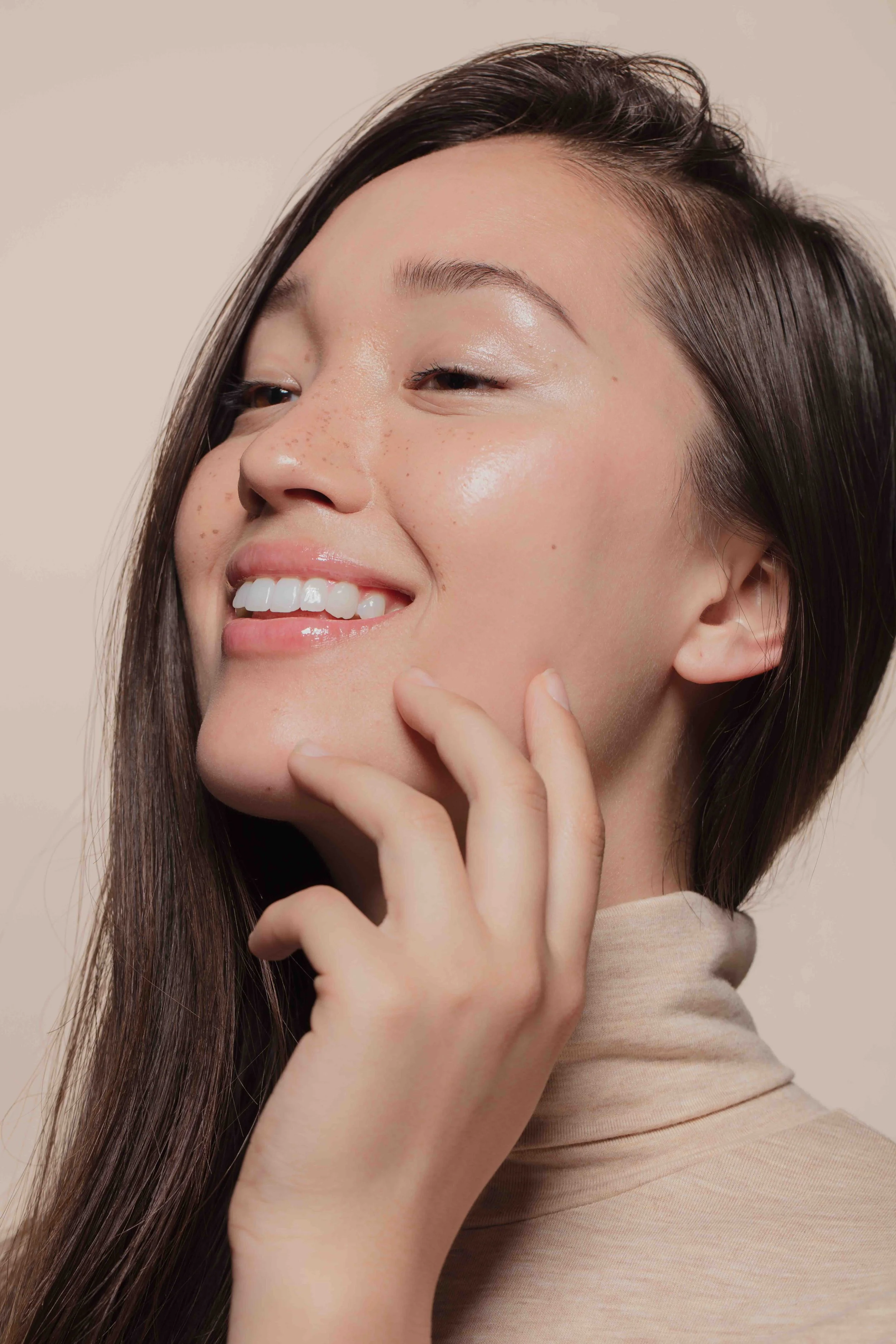 Close-up of a smiling woman with long dark hair, showing freckles, smooth skin, and wearing a beige turtleneck.
