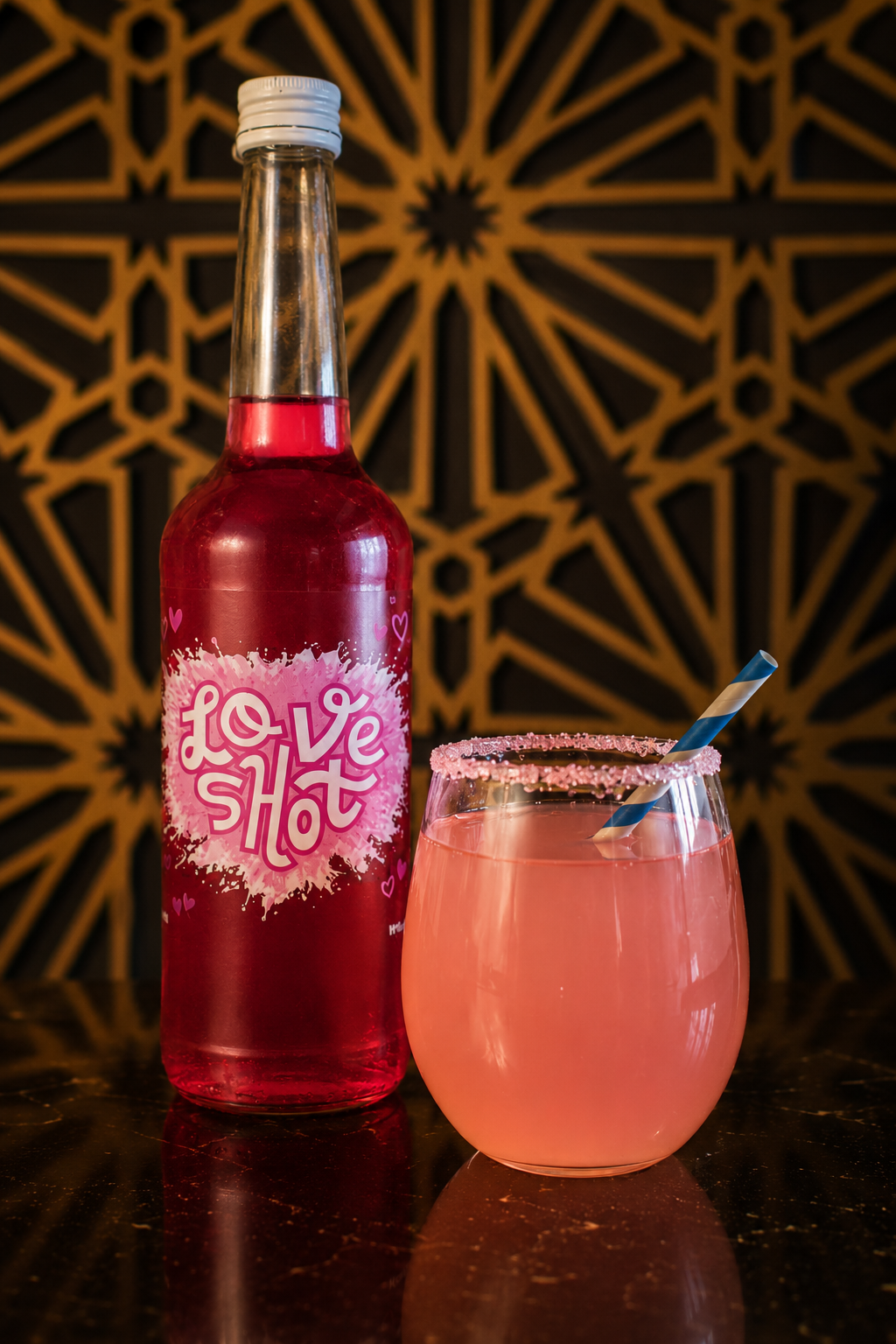 Een fles met een roze label dat zegt 'Love Shot' staat naast een glas gevuld met een roze drank en een rietje, op een donkere tafel met een decoratieve achtergrond.