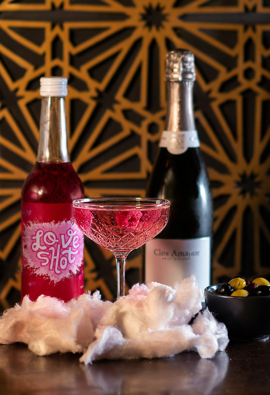 Een glas roze champagne, een fles champagne, een fles met de label 'Clos Amor', een zwarte kom met olijven, een bord met roze en zwarte olijven, en wat roze en witte zachte stof rondom, op een donkere tafel met een decoratieve achtergrond.
