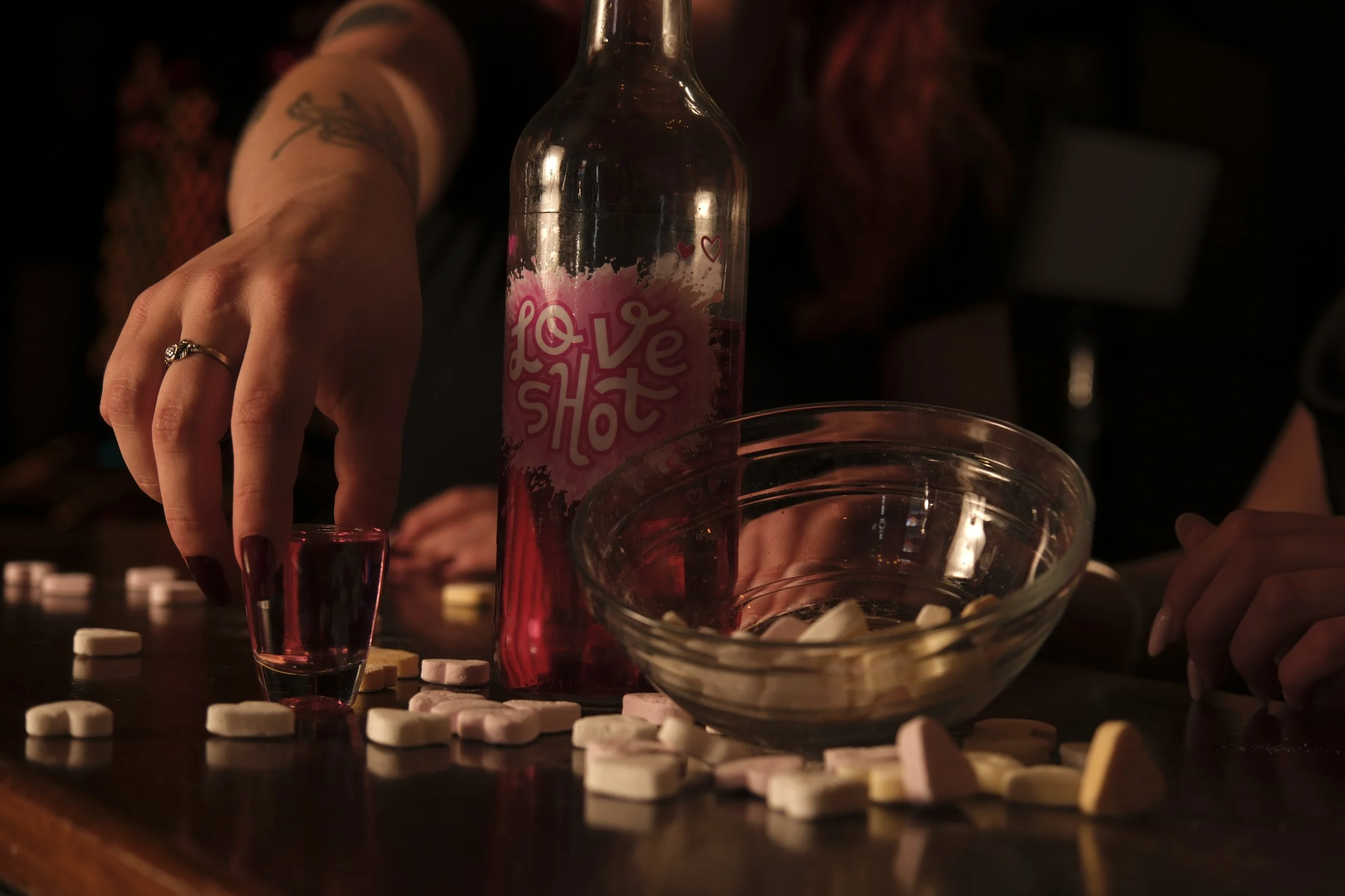 Een persoon met een ring op de vinger giet een roze drankje in een klein glas, op een donkerkleurige tafel met met witte en gele tabletten en een grote glazen kom