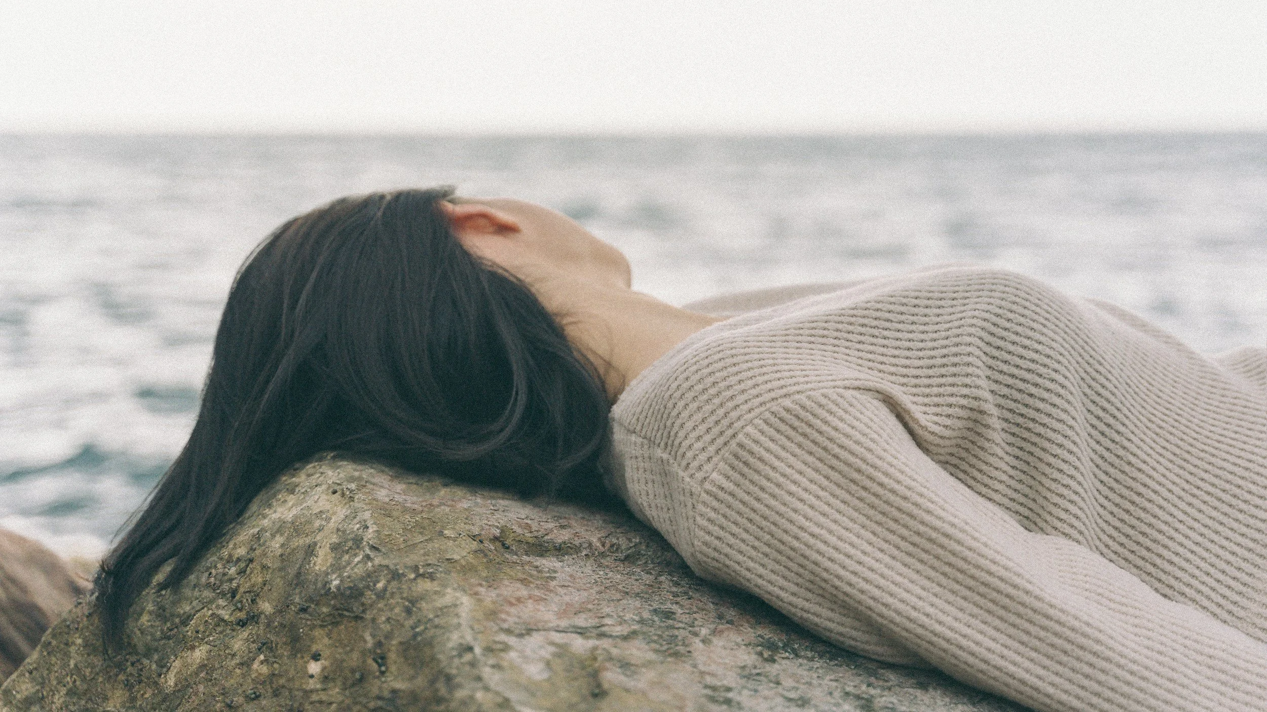 A woman with long dark hair lying on a large rock by the ocean, wearing a beige knitted sweater, with her face turned away and eyes closed.
