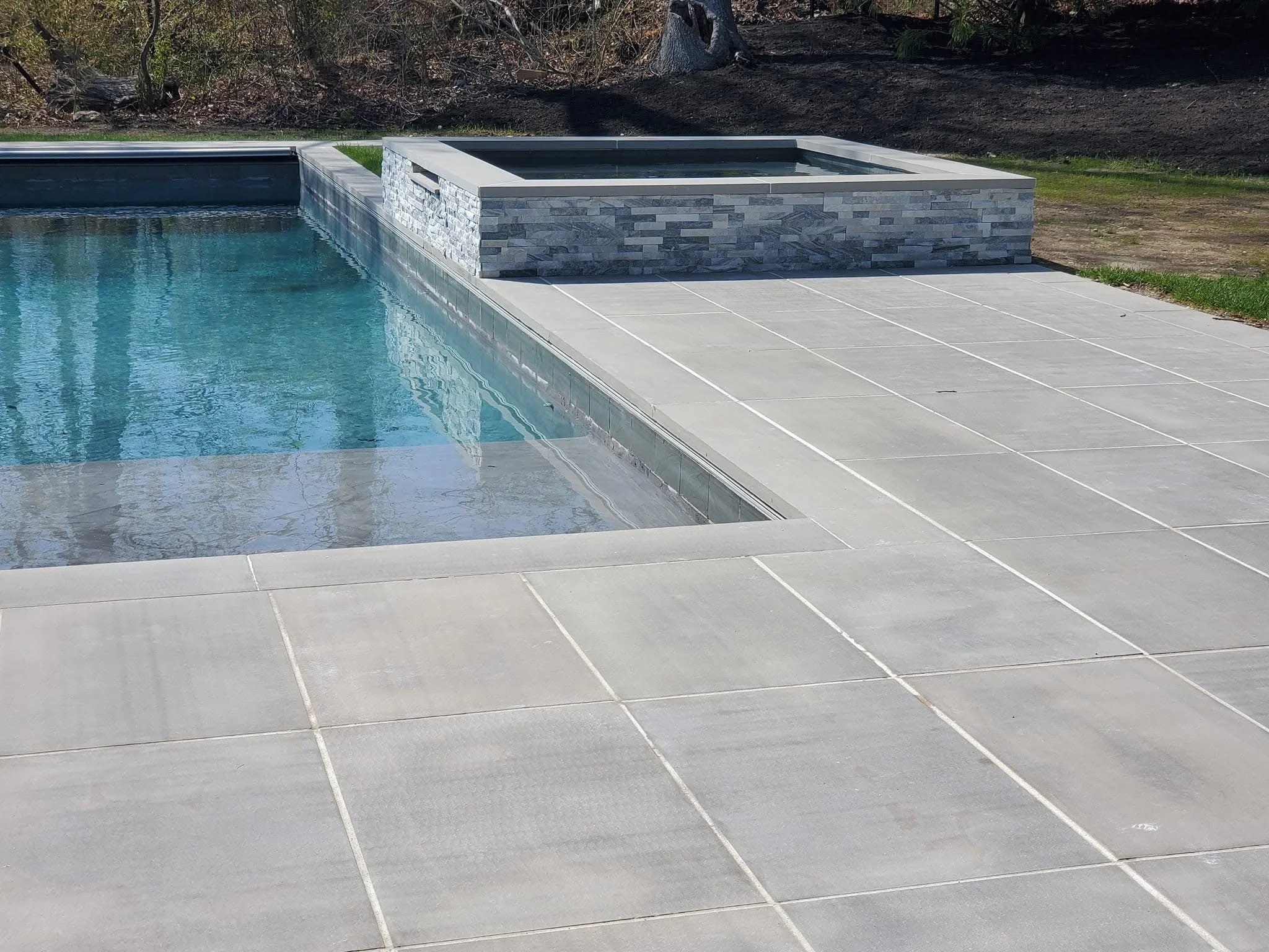 A backyard swimming pool with a raised hot tub made of gray stone on the side, surrounded by light-colored tile decking and a grassy area with some trees and dirt in the background.