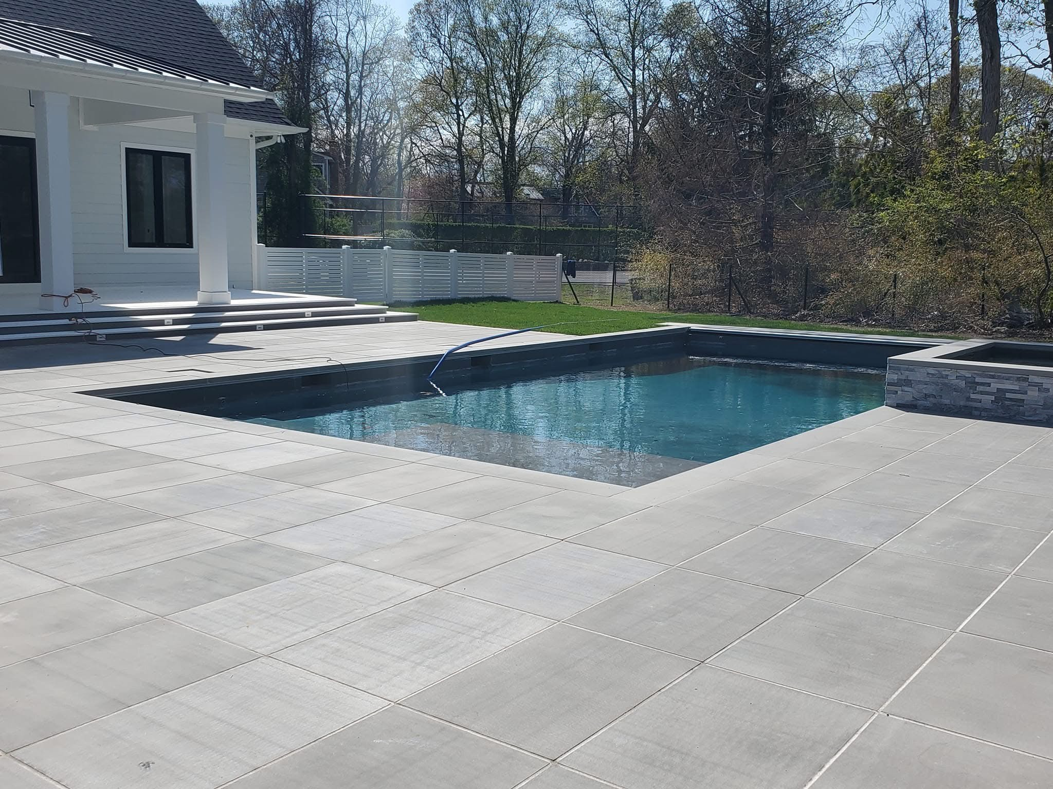 Backyard with swimming pool, white house with porch, trees in the background, and a clear sky.