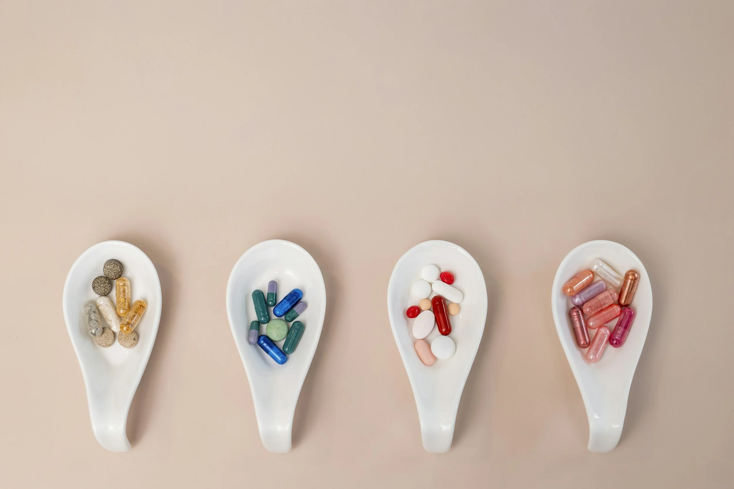 Quatre cuillères en porcelaine blanche contenant différentes pilules et compléments alimentaires de différentes formes, couleurs et tailles, disposées en ligne sur une surface beige.