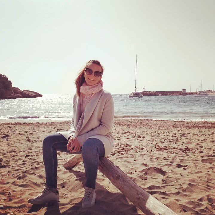 Une femme souriante portant des lunettes de soleil, un manteau, une écharpe et des chaussures d'hiver, assise sur un vieux tronçon de bois sur une plage, avec la mer et un bateau en arrière-plan, par une journée ensoleillée.