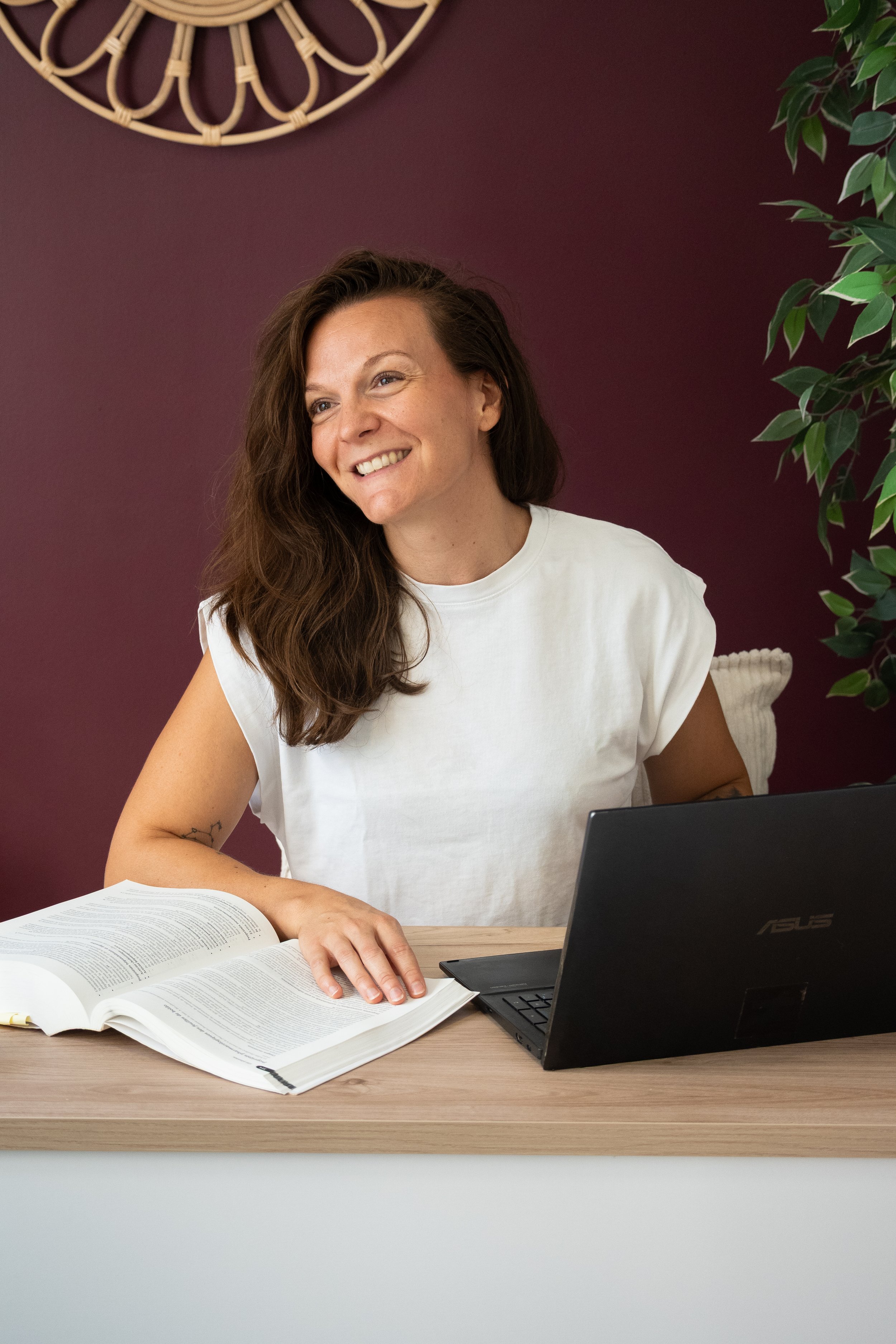 Une femme souriante avec des cheveux bruns longs, portant un t-shirt blanc, assise à un bureau avec un ordinateur portable Asus ouvert devant elle, un livre ouvert, et un carnet, dans une pièce avec un mur violet et une plante verte à droite.