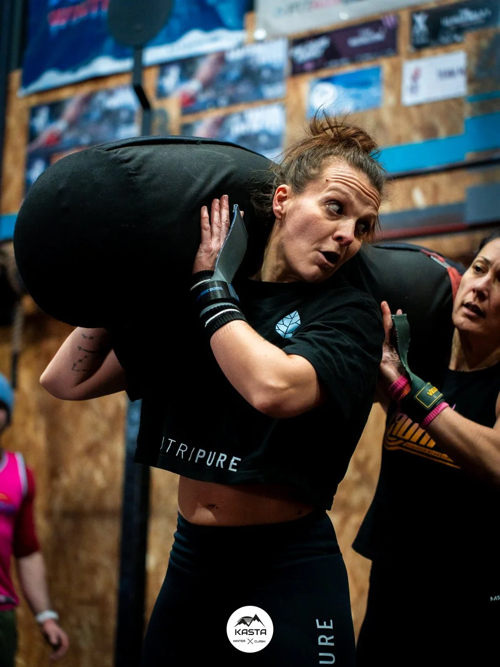 Athlète féminine de CrossFit portant un lourd sac lors d’un entraînement, encouragée par une coéquipière.