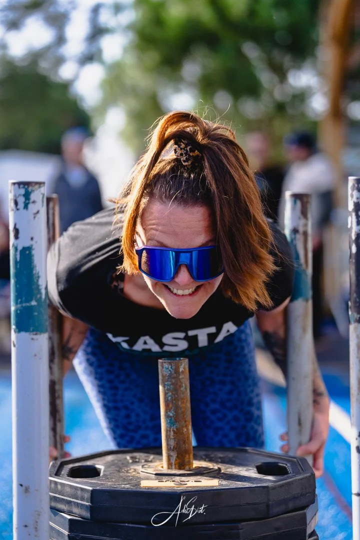 Une femme souriante portant des lunettes de soleil réfléchissantes tente de soulever une lourde plateforme lors d'une activité en plein air, avec d'autres personnes floues en arrière-plan.
