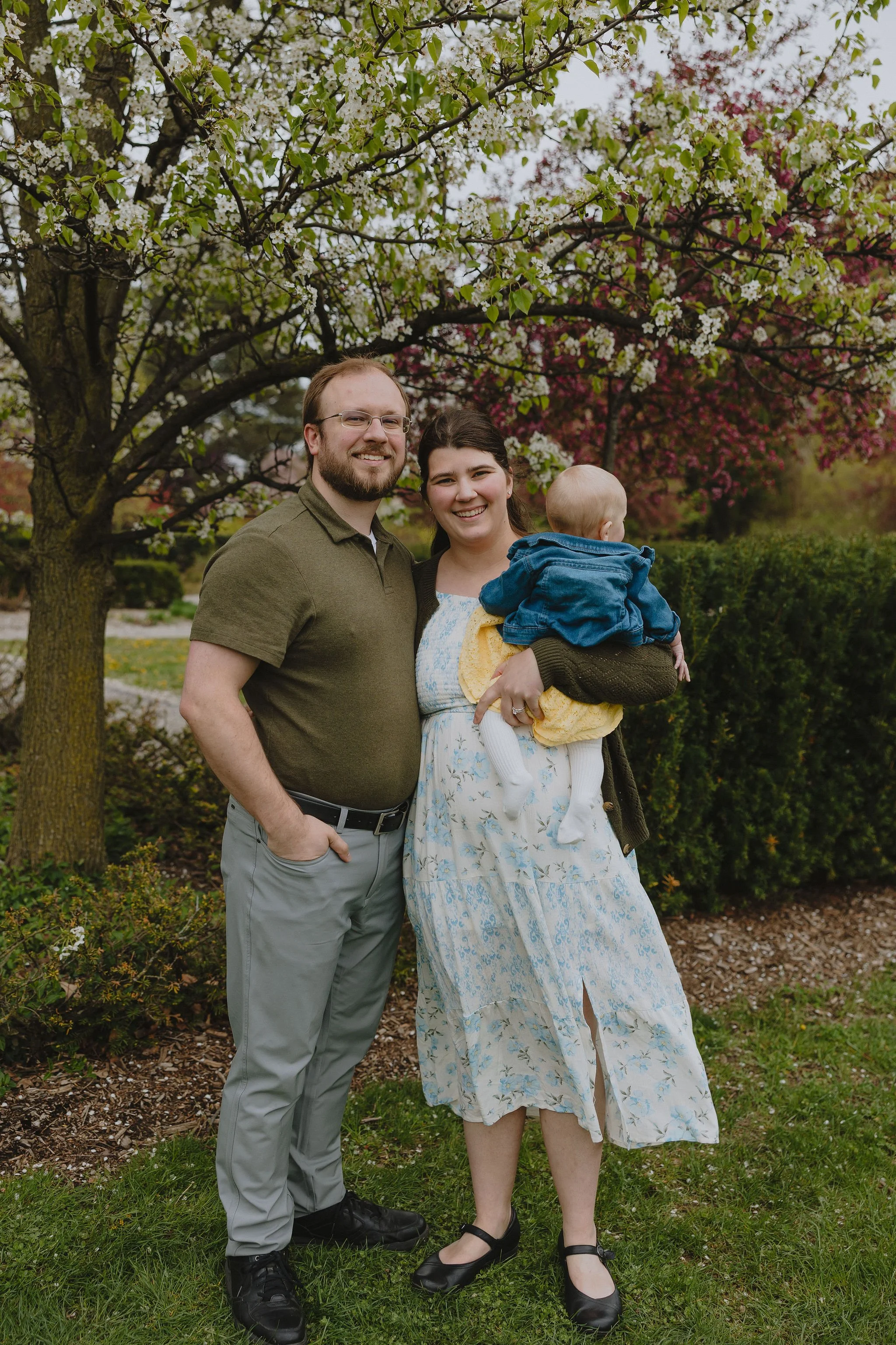 A family of three standing outdoors in front of blooming trees. The man has glasses and a beard, wearing a green polo shirt and gray pants. The woman has dark hair and is holding a baby, wearing a long floral dress. The baby has a blonde head and is dressed in a blue jacket and yellow dress. They are smiling and standing on green grass and mulch, with flowering trees in the background.