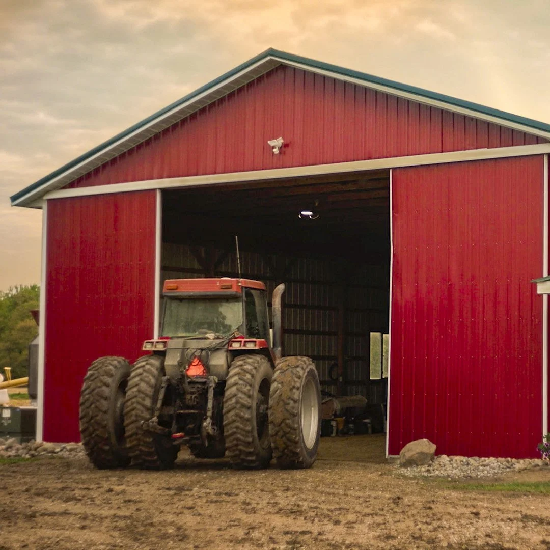A still frame from a commercial of a red tractor driving into a red barn during sunset.