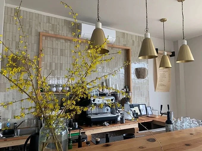 A bar or cafe counter with a large yellow flowering branch in a glass vase, pendant lights overhead, coffee equipment, framed photos, and glasses on the counter.