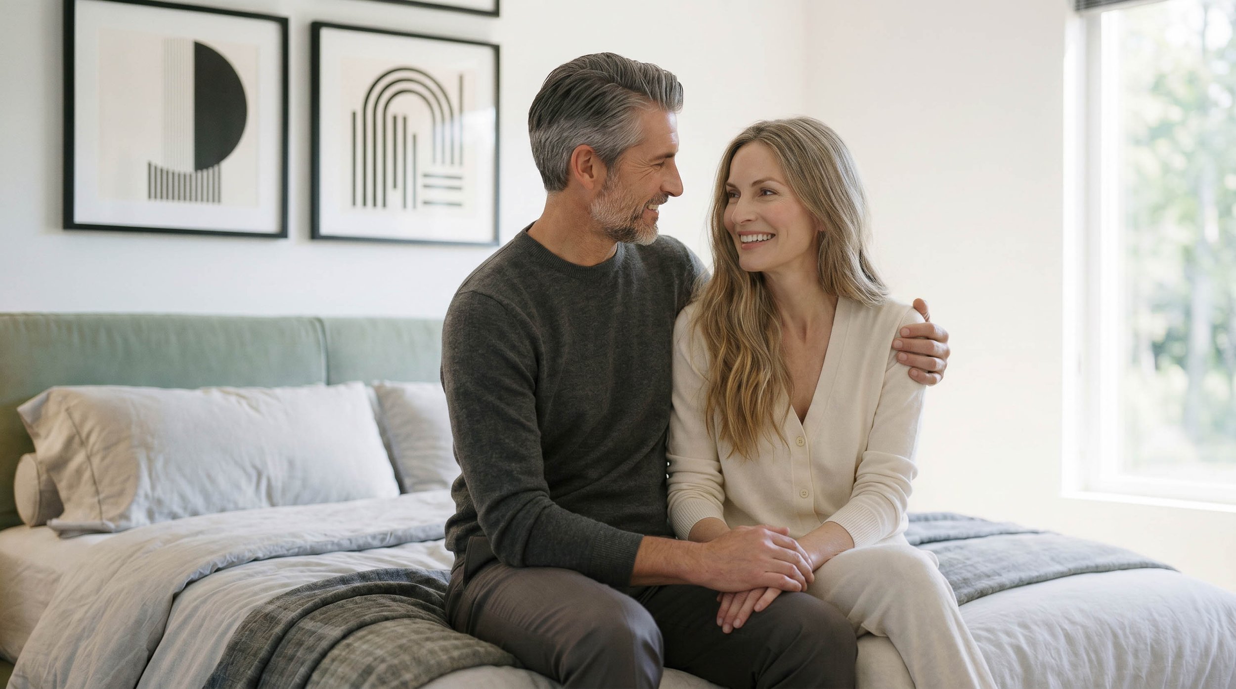 A middle-aged man and young woman sitting close together on a bed, smiling and looking into each other's eyes in a bright, modern bedroom.