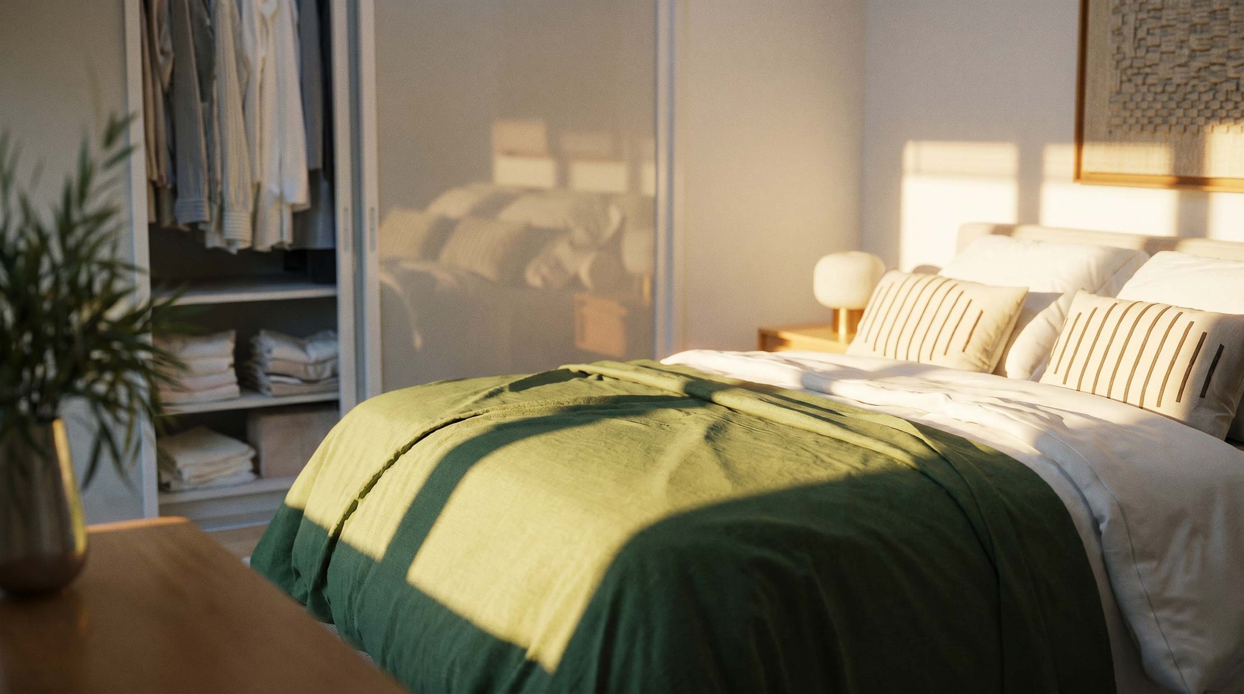 A neatly made bed with white pillows and a green blanket in a sunlit bedroom. There is a bedside table with a lamp and a framed artwork on the wall. An open closet with hanging clothes and folded linens is visible in the background.