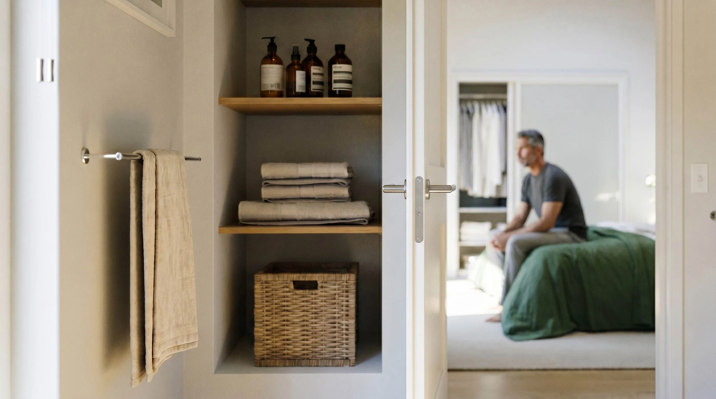 An open closet with shelves holding bottles, towels, and baskets, with a man sitting on a bed in the background.