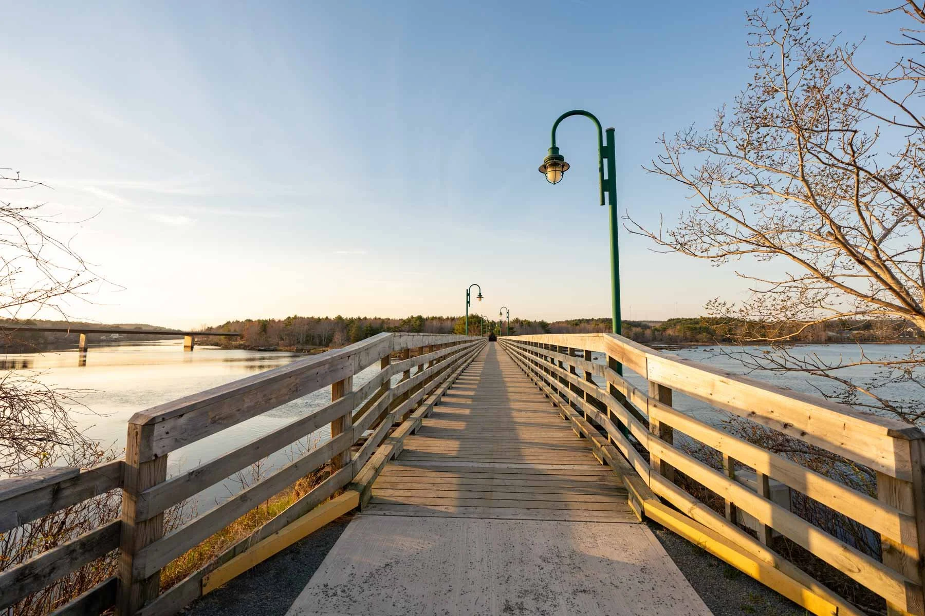 Wooden bridge over a body of water with a railing on each side, green street lamps, and leafless trees in the background during sunset or sunrise.