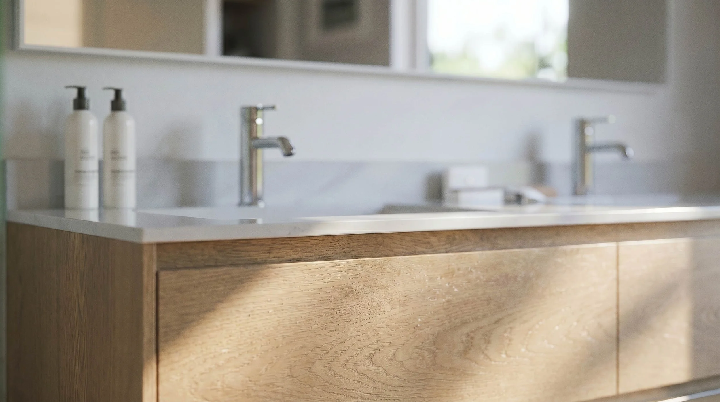 Close-up of a bathroom with a wooden vanity, a white countertop, two modern silver faucets, and two white soap dispensers.