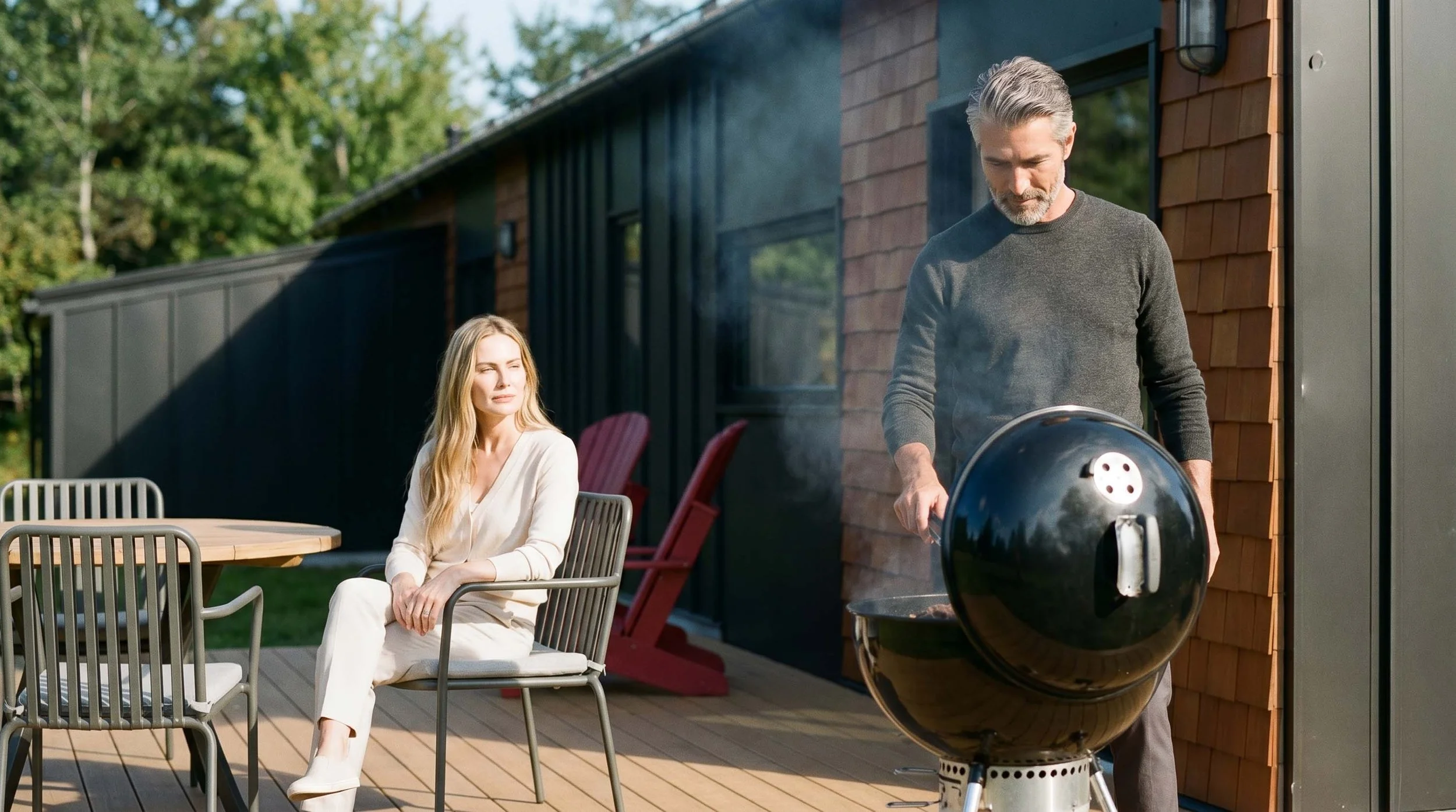 A man is grilling on a backyard patio while a woman sits nearby on a chair, outdoors with a modern house and trees in the background.