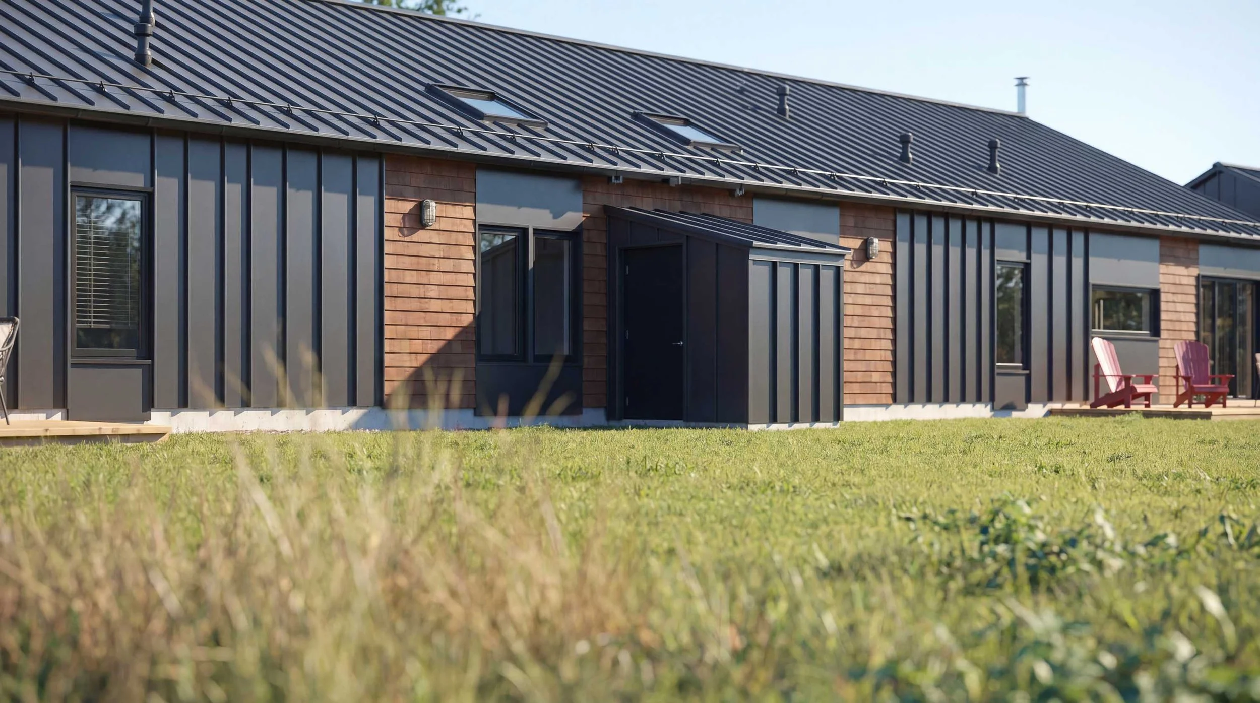 Modern house with dark metal and wooden siding, multiple skylights, and an outdoor grass yard with two red chairs.
