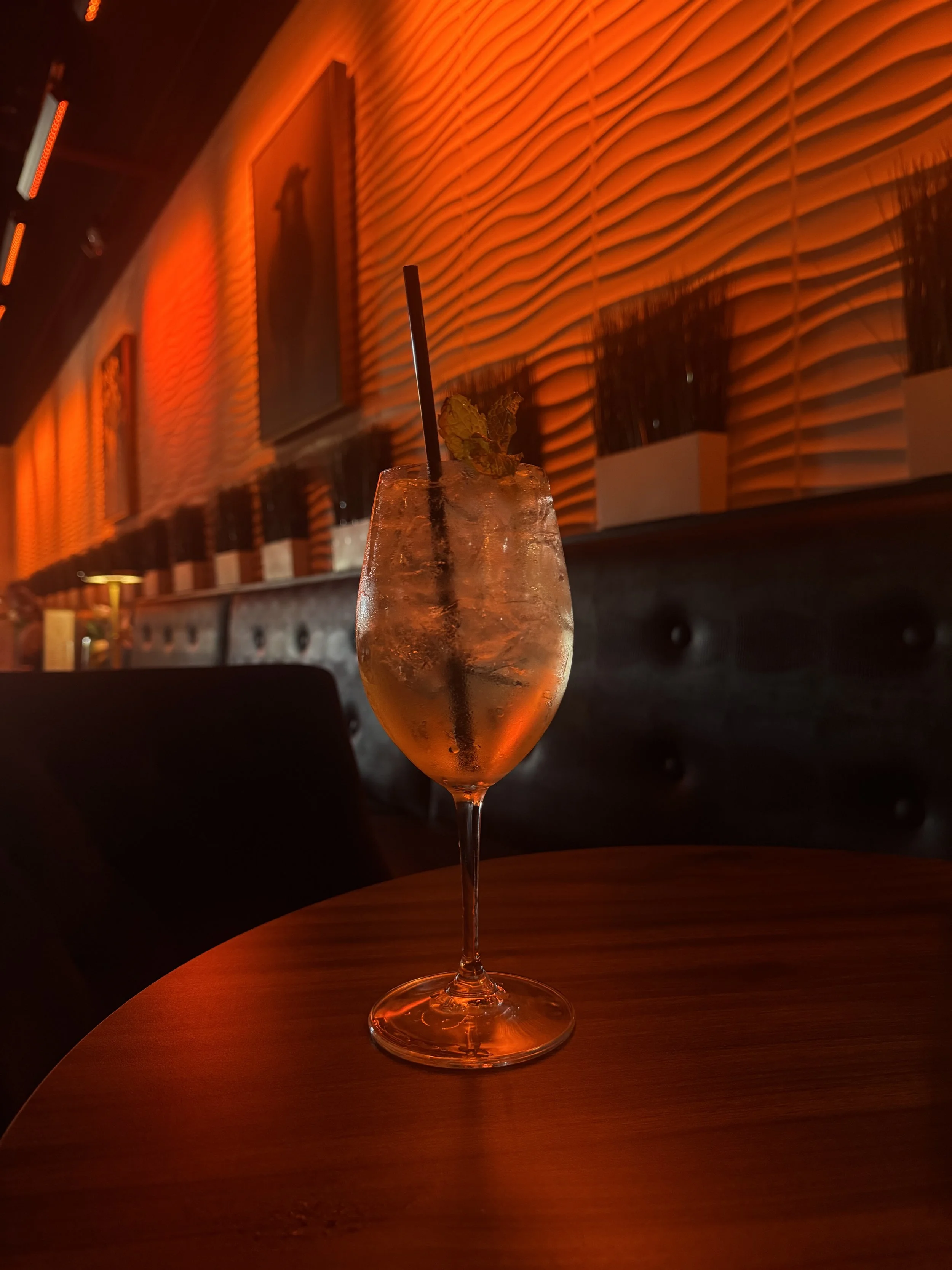 A tall glass with a cocktail garnished with a mint leaf and a black straw, set on a round wooden table in a dimly lit, stylish lounge with orange-red lighting and textured wall panels in the background.
