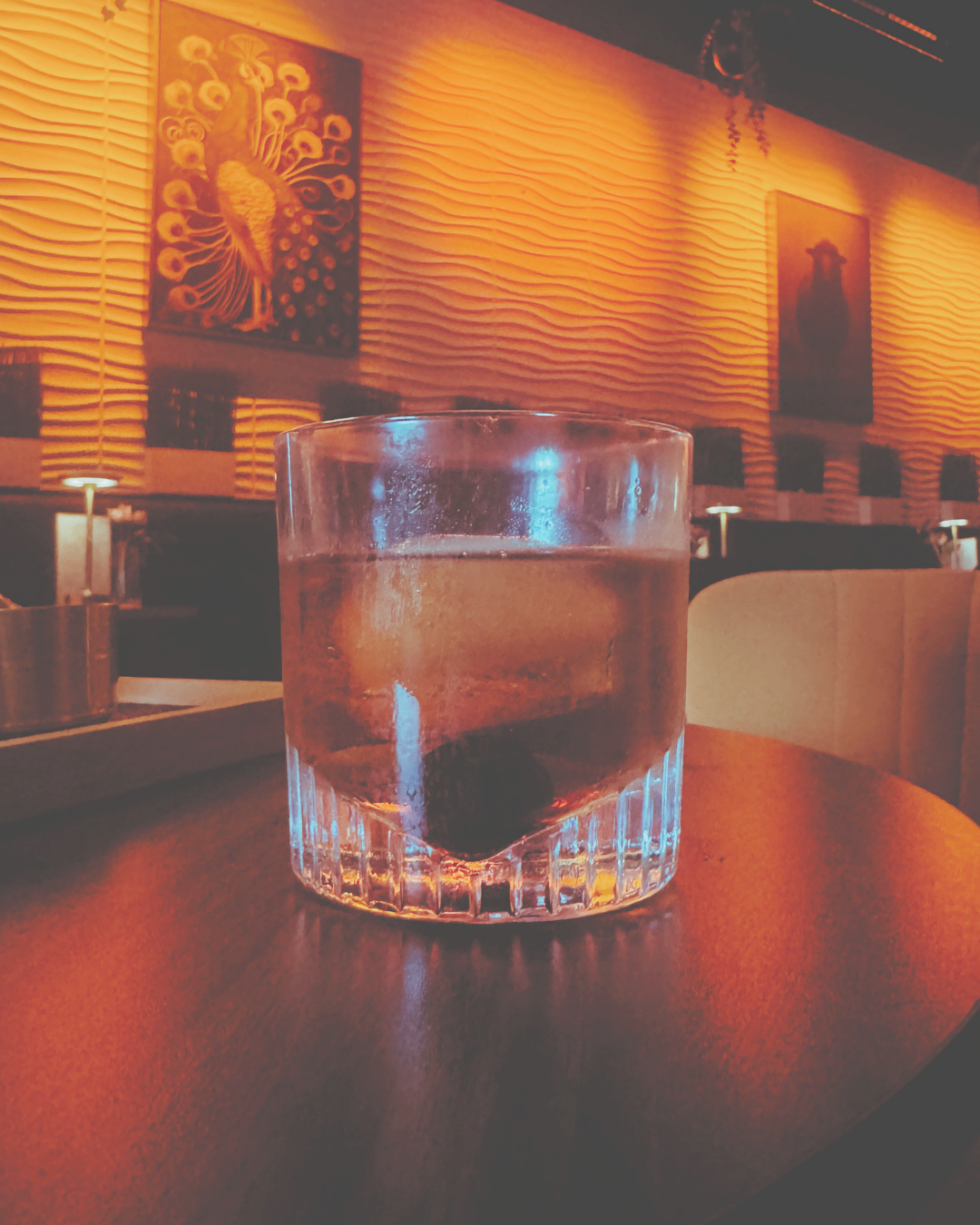 A glass of whiskey with a cherry at the bottom on a wooden table in a dimly lit lounge or bar with artwork of a peacock and a dog on the textured wall in the background.