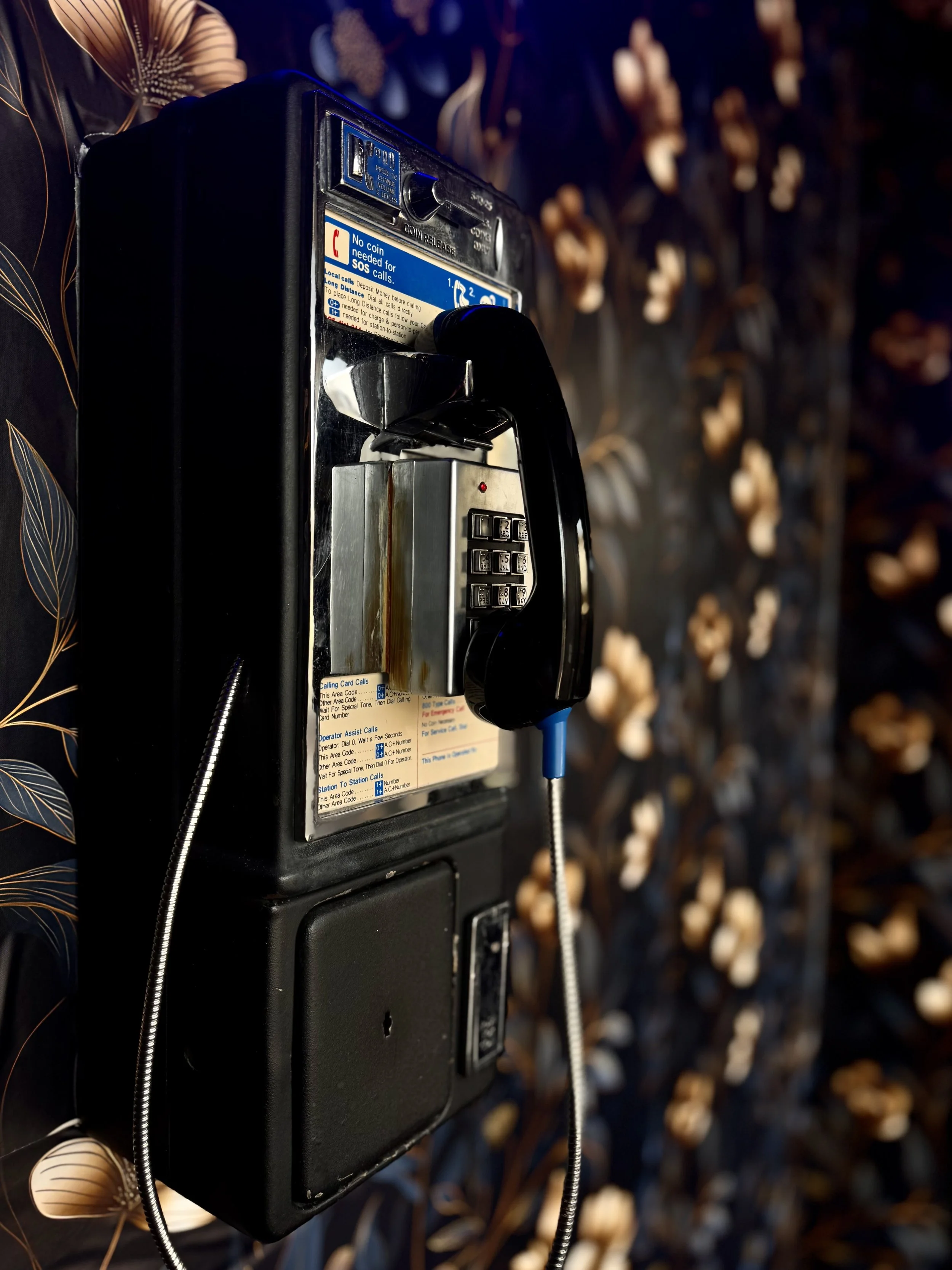 An old payphone mounted on a wall with a background of carved wood or shell art. The payphone has a black plastic exterior, a metal keypad, and a corded handset hanging on the side.
