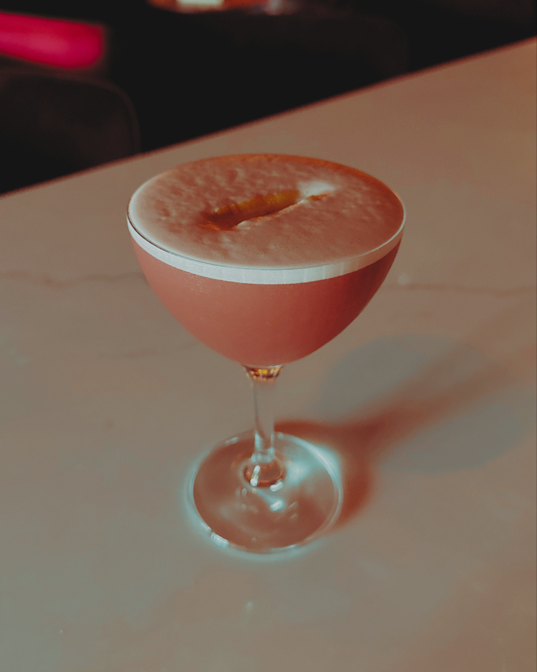 A pink cocktail in a coupe glass with foam on top, garnished with a small lemon peel.