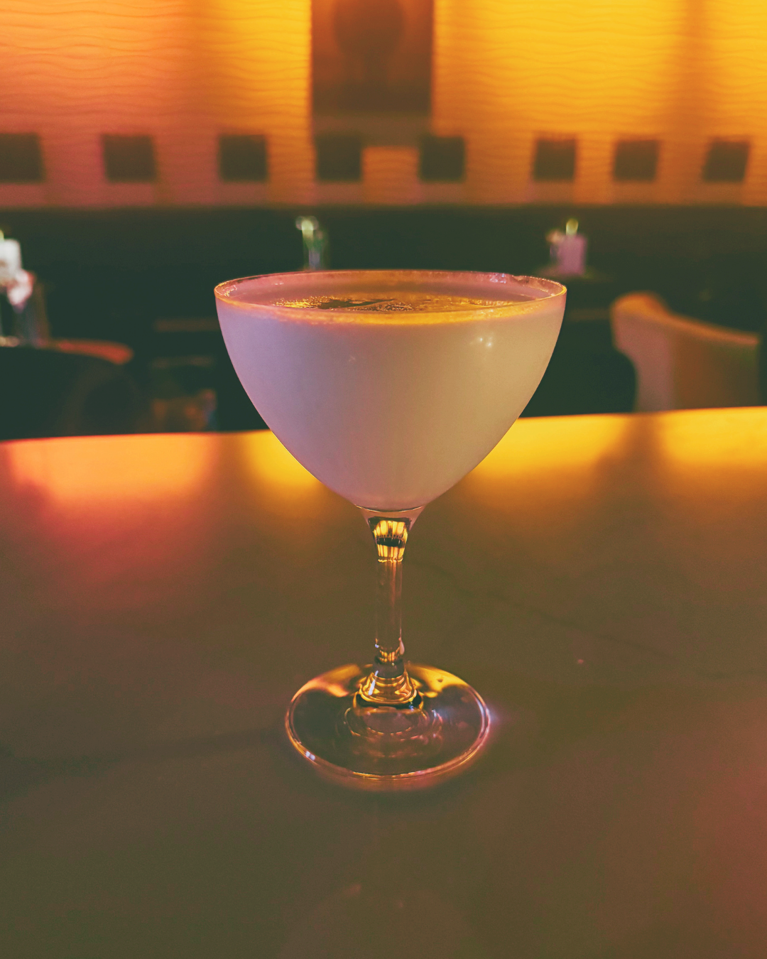A cocktail glass filled with a pale yellow drink on a bar counter with warm, colorful lighting and blurred background seating.