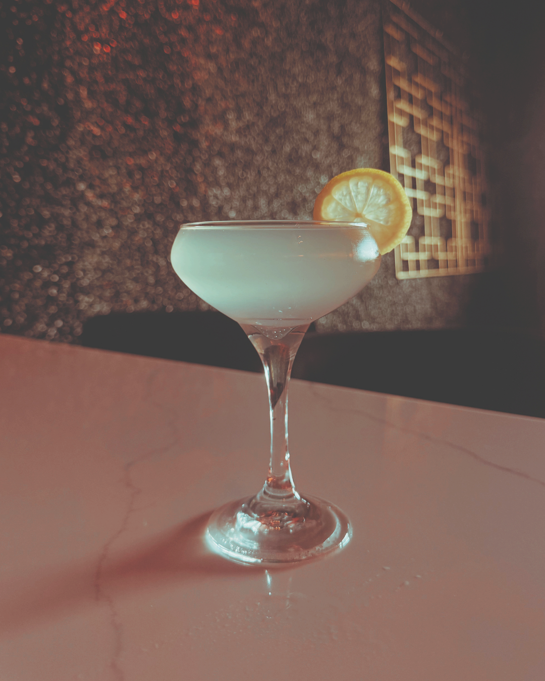 A cocktail glass with a lemon slice garnish on the rim, filled with a light-colored drink, placed on a marble bar counter with a textured, dark wall and a menu board in the background.