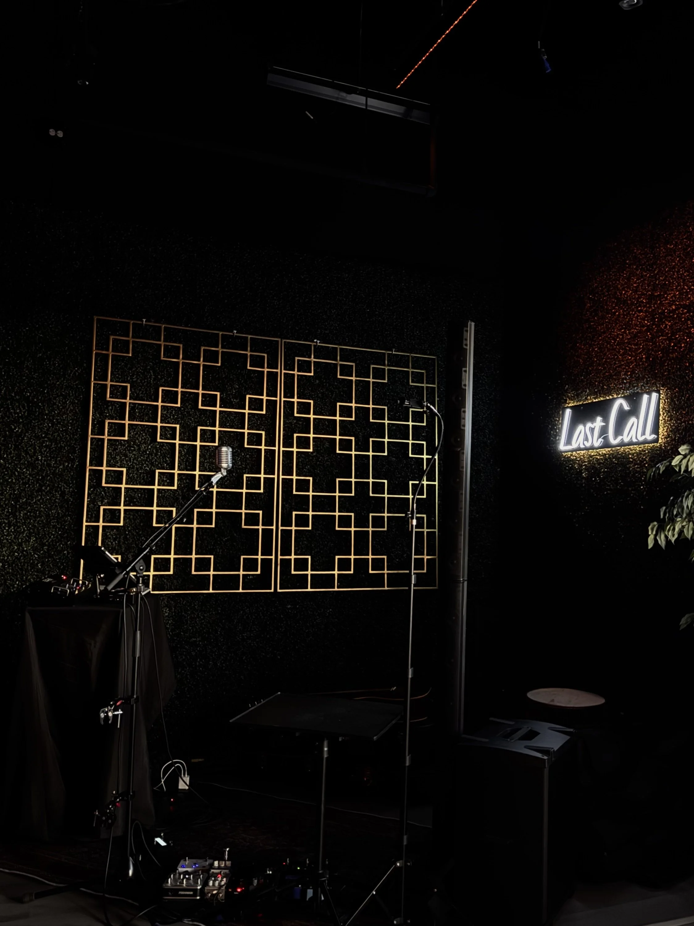 A dimly lit stage area with a neon sign that reads "Last Call" on the right side. There are two mounted microphones on stands in front of a geometric wall decoration, and various audio equipment on the floor.